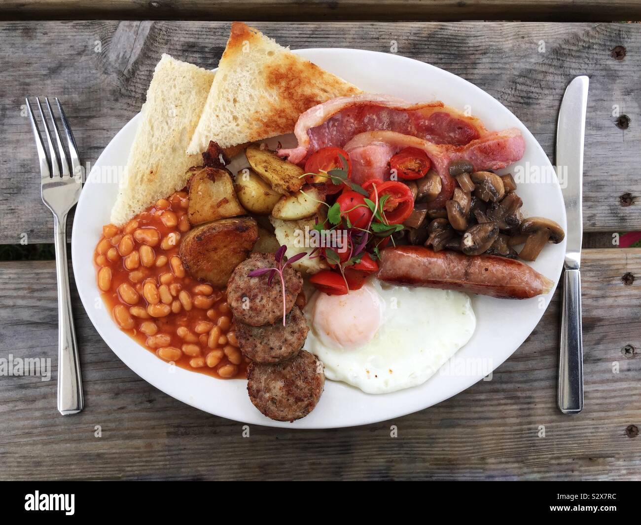 Looking down from above onto a plate full of full English breakfast on the wooden table with knife and fork Stock Photo