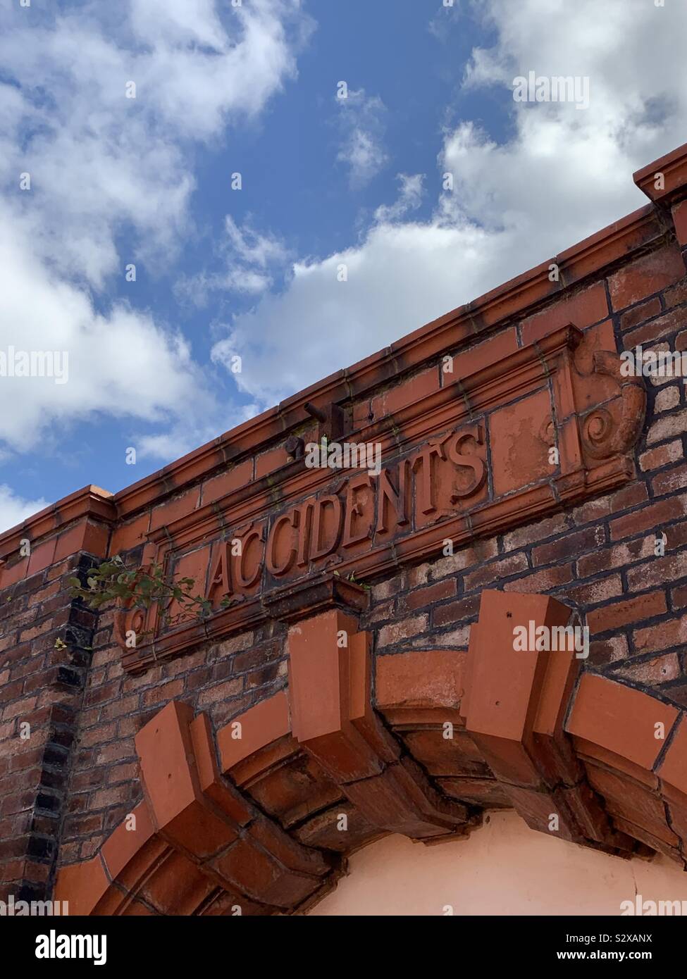 Victorian entrance archway saying accidents with old weed filled brickwork against a blue cloud sky Stock Photo