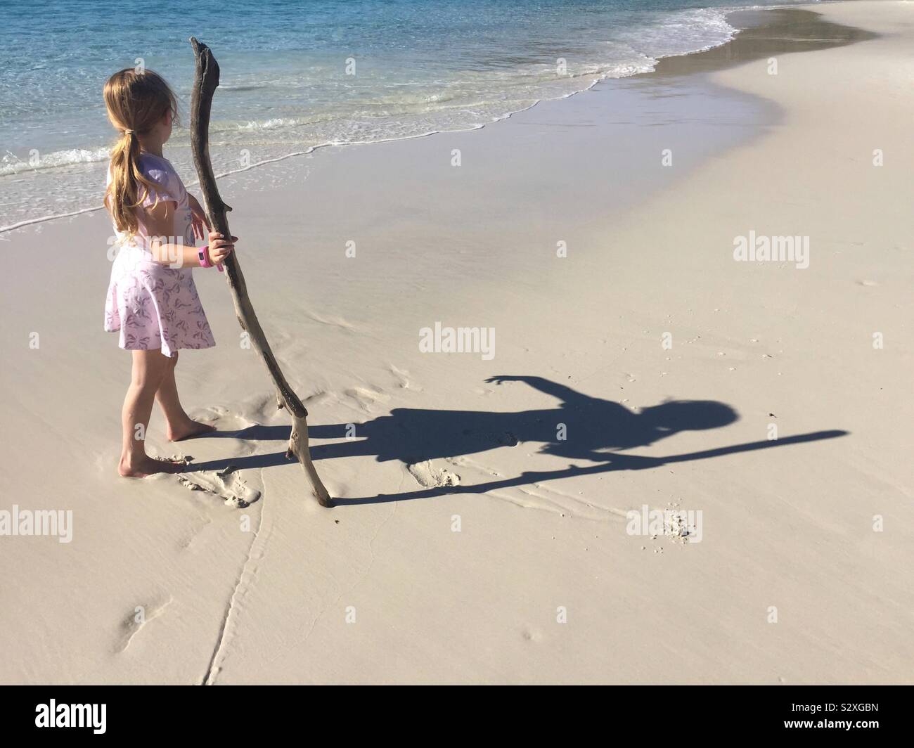 Young girl in pink summer dress plays on beach on bright sunny day. Girl having fun, playing and exploring ocean beach. Concept vacations, childhood, exploration and lifestyle. Stock Photo