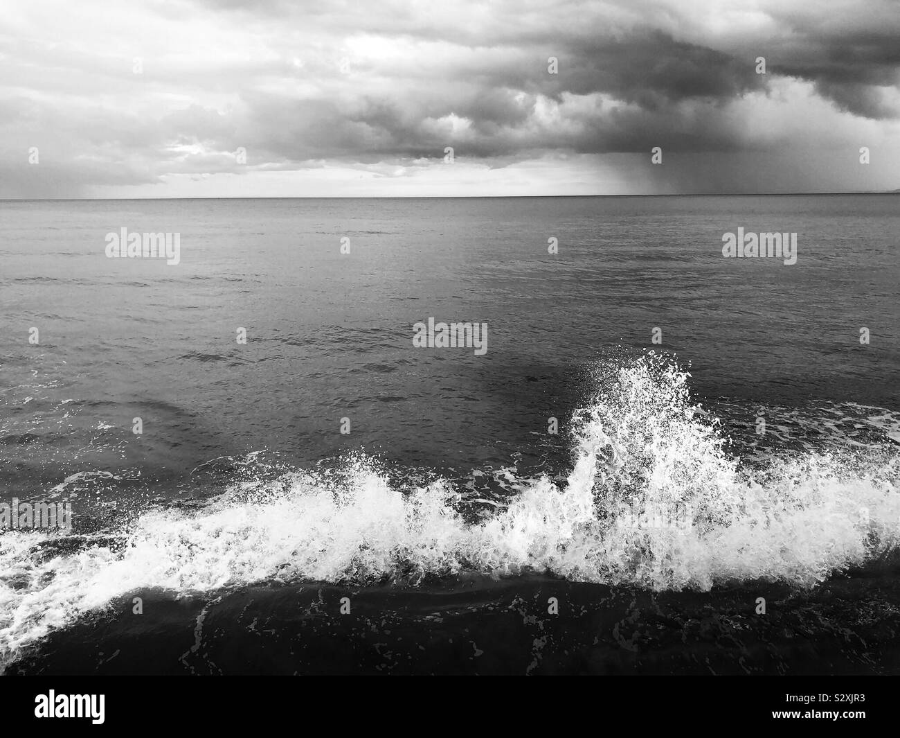 Black and white image of a breaking wave at high tide with rain falling in the distance Stock Photo