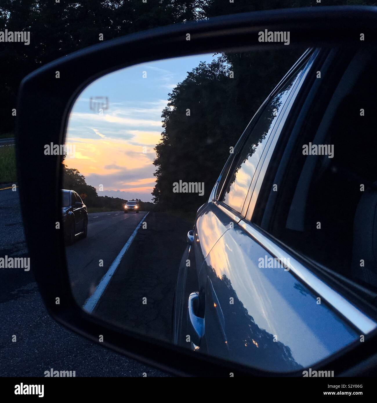 Road trip autumnal sunset reflected in car mirror intense vivid beautiful colors colours and reflections in glass and side of automobile with cars traveling on country road. Stock Photo