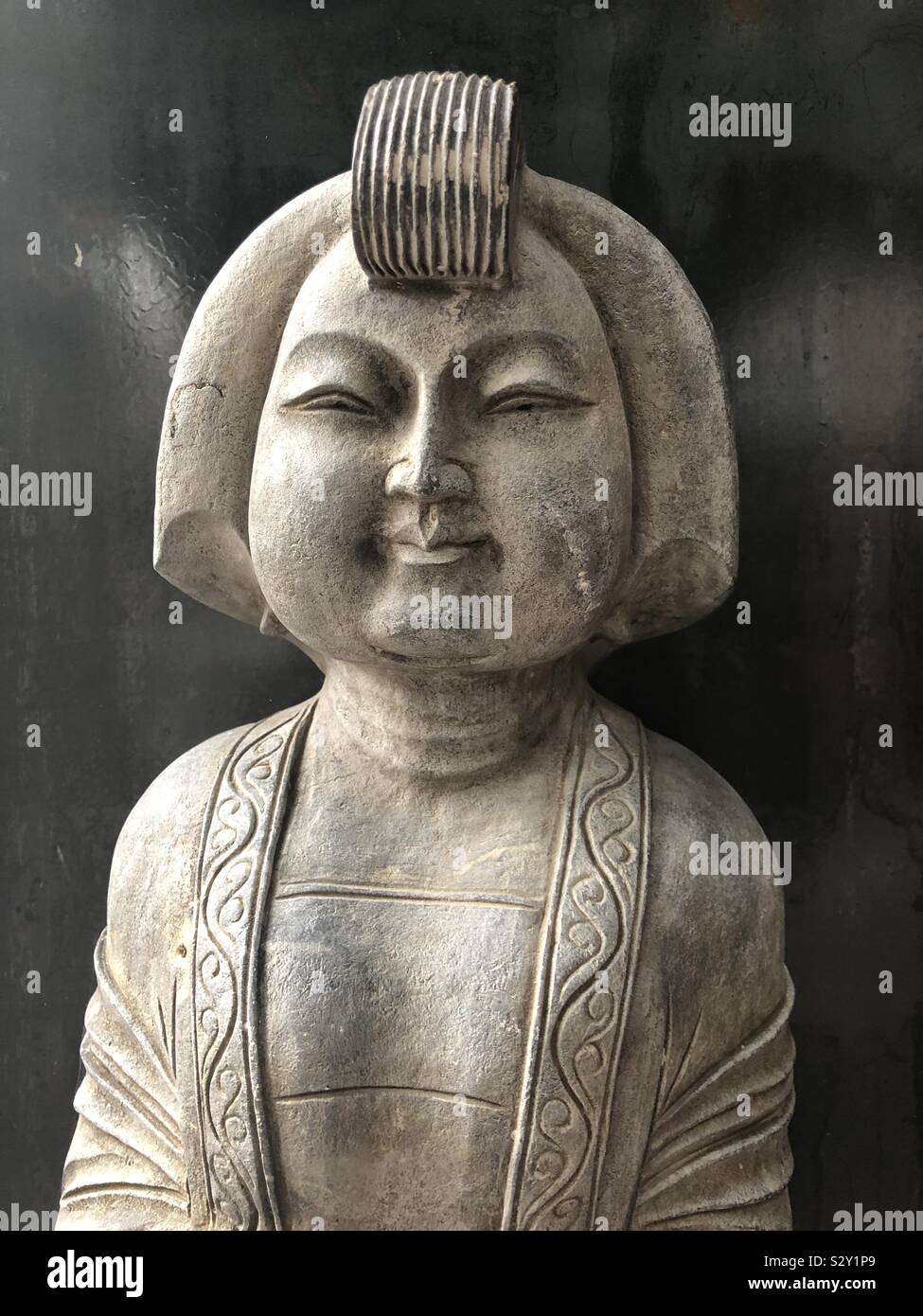 Frontal view of the upper half of a stone statue featuring a round Asian face, bobbed hairstyle with quiff against a black background. Stock Photo