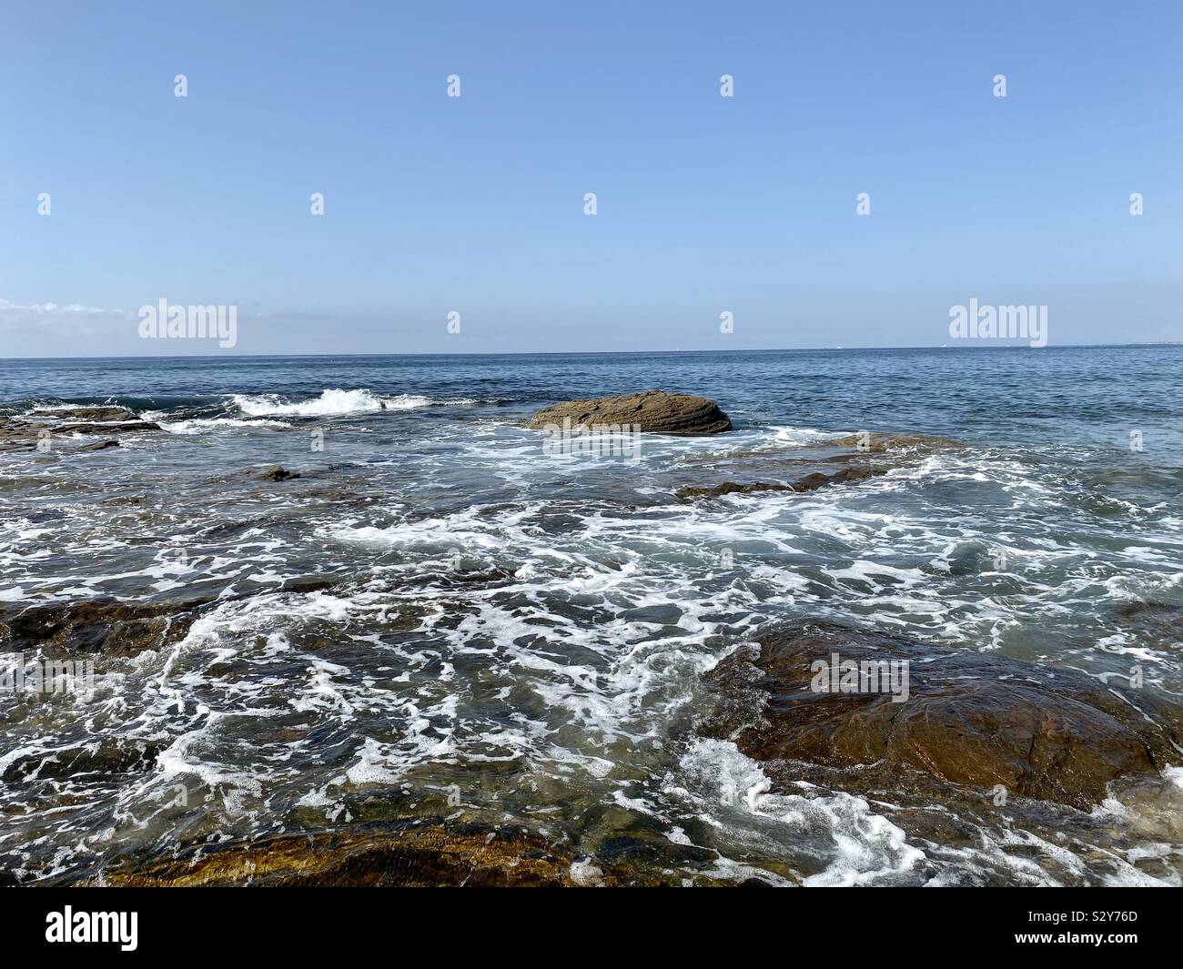Seascape of Pacific Ocean water with waves flowing over rock formations and blue skies - Smartphone Captured Stock Image