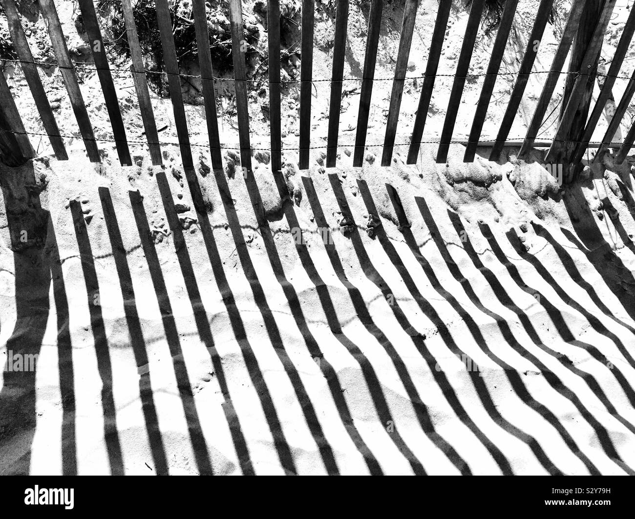 A wooden picket fence along a sandy footpath leading to the beach has its shadow forming an abstract pattern in the sand. Stock Photo