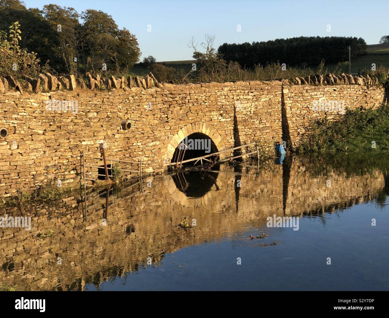 A bridge over a flooded Cotswold stream in beautiful late afternoon sunshine, highlighting the mellow honey coloured Cotswold stone, a peaceful Autumn scene. - Smartphone Captured Stock Image