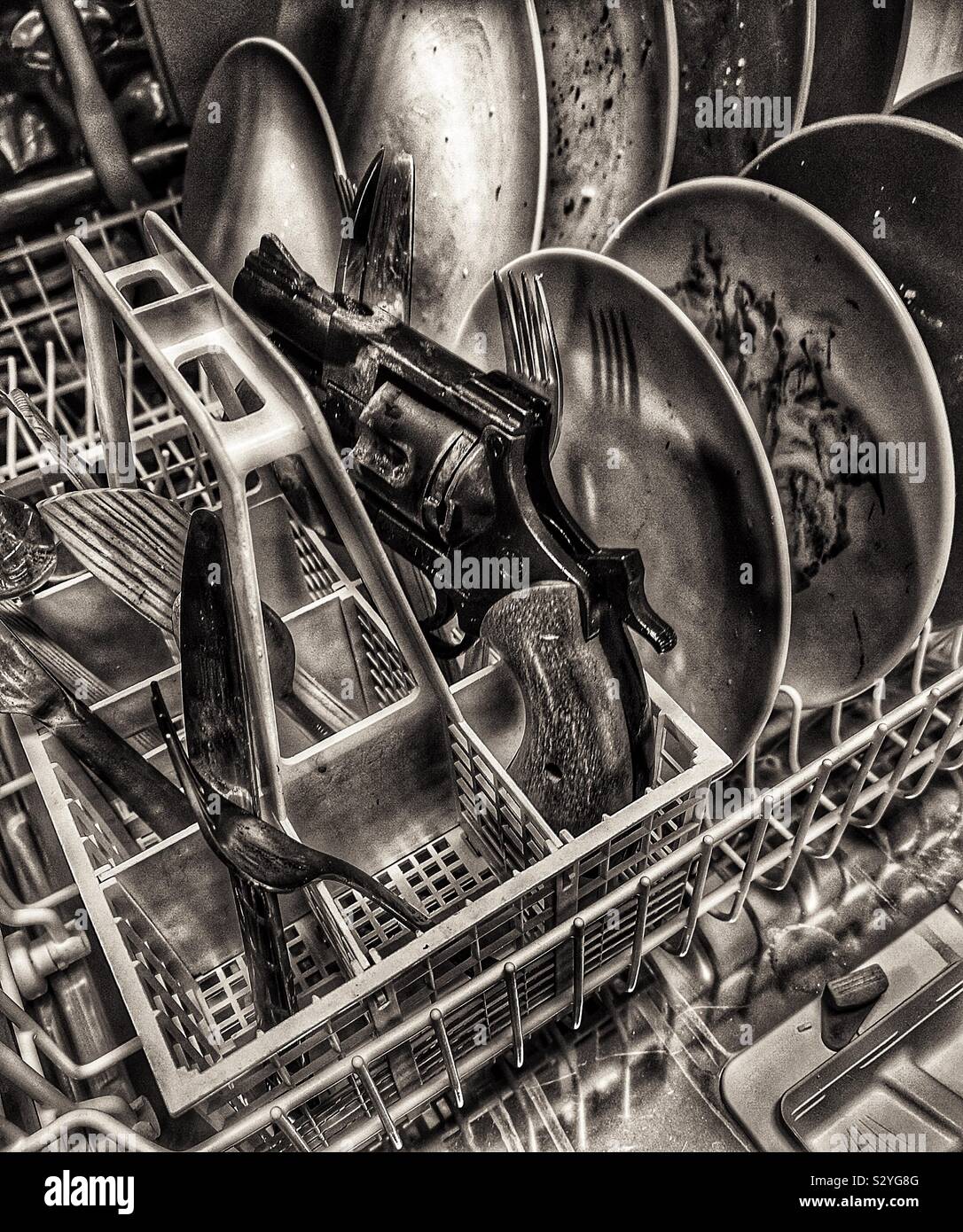 Black and white image of pistol in dishwasher amongst plates and cutlery - Smartphone Captured Stock Image