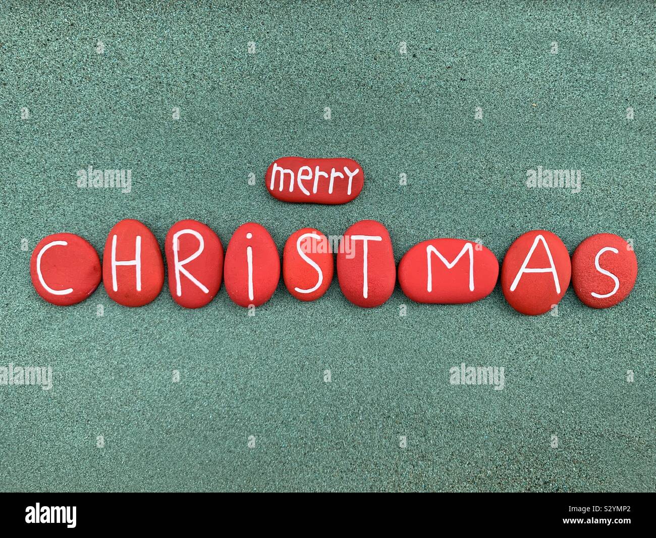 Merry Christmas text with red colored stones over green sand - Smartphone Captured Stock Image