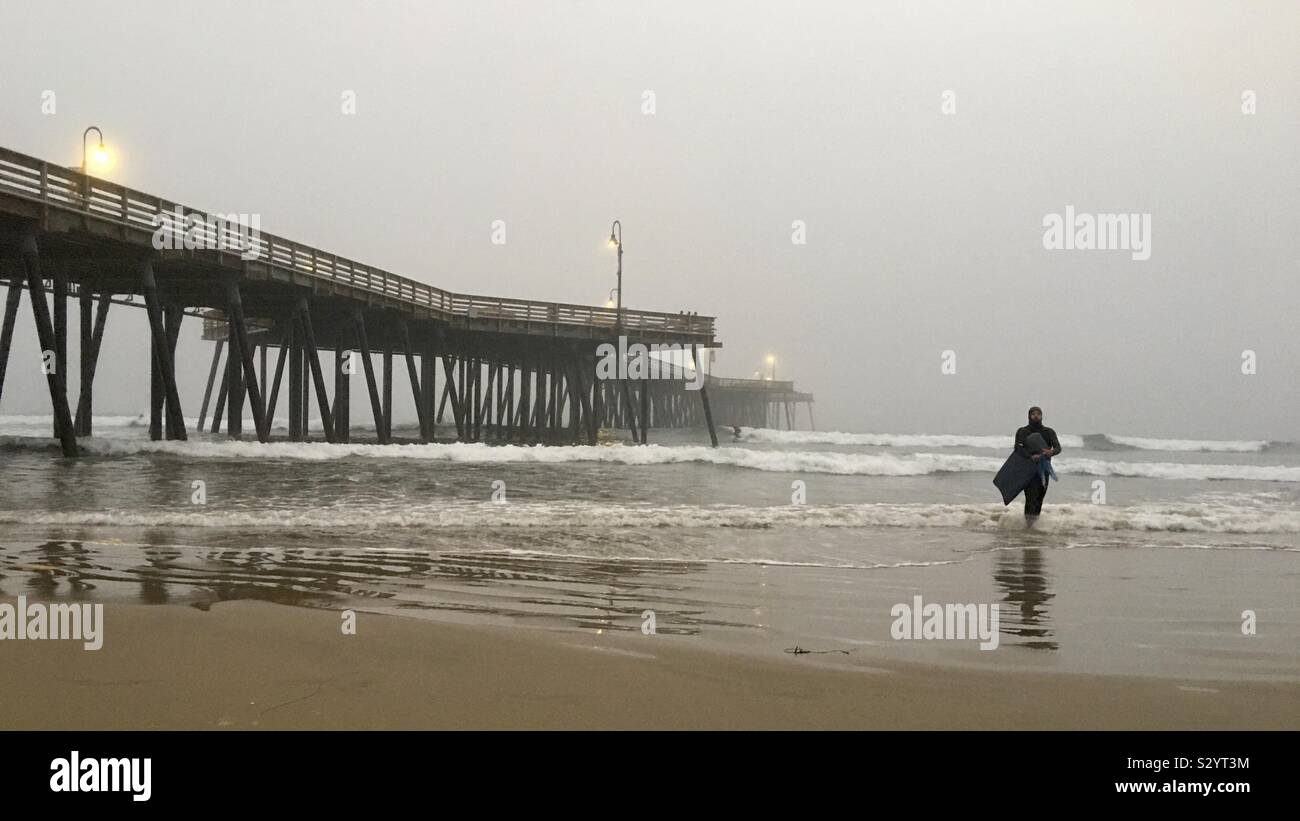 PISMO BEACH, CA, OCT 2019: surfer in black wetsuit walks out of Pacific Ocean next to the pier, on a foggy late afternoon Stock Photo
