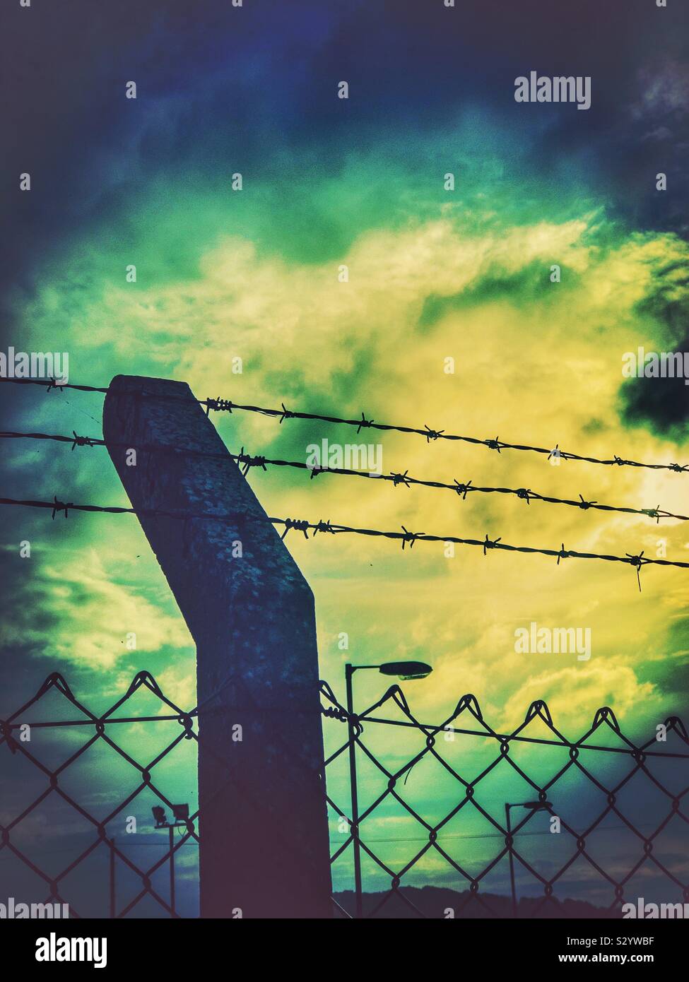 Concrete post with barbed wire and mesh fence against a dramatic cloudy sky. Stock Photo