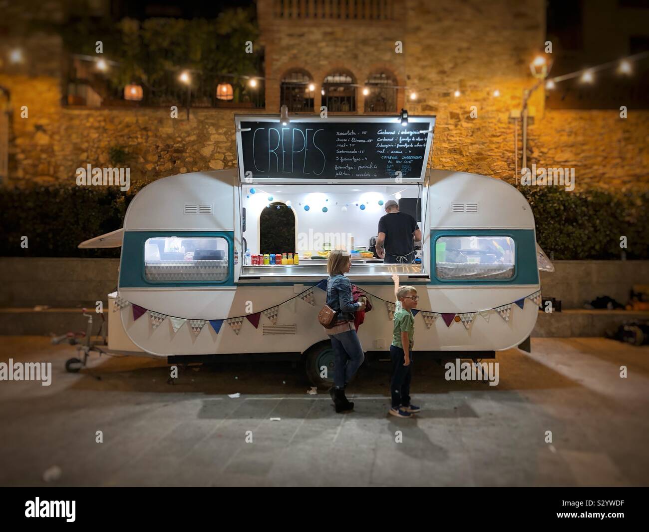 Mother and son waiting for crepes at a retro styled fast food trailer kiosk in the centre of Begur, Spain, late evening. - Smartphone Captured Stock Image