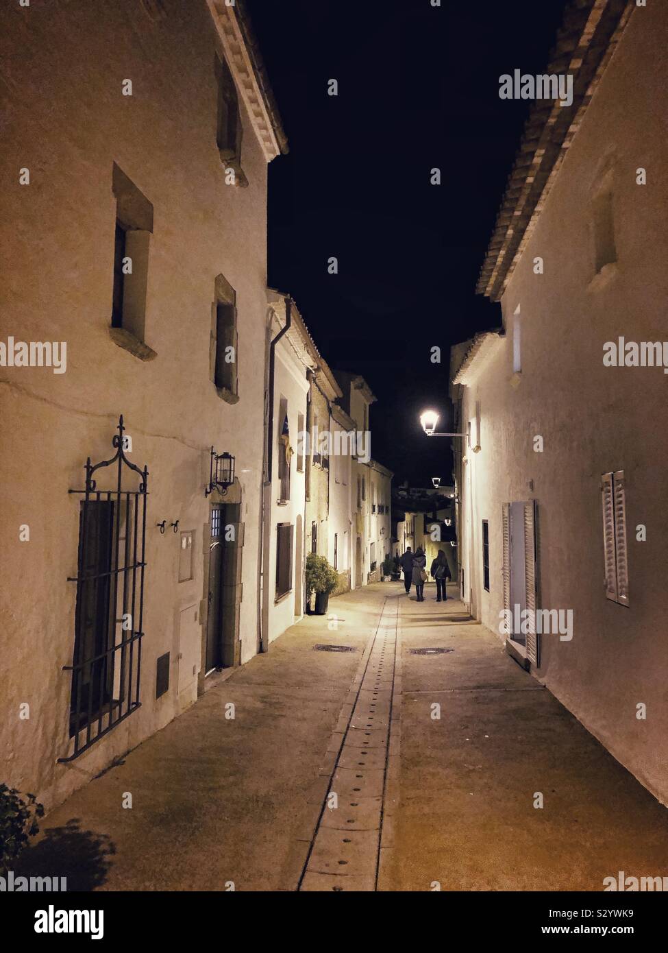 A typical street in the old town of Begur, Spain, at nighttime with three people under a streetlight at the bottom of the road. - Smartphone Captured Stock Image
