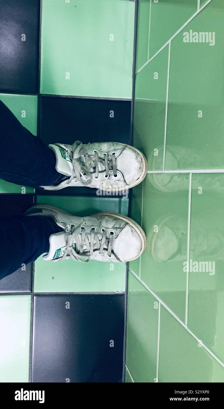 Girl sat down on the toilet seat in a hostel in Europe. Trainers on a green and black tiled floor. - Smartphone Captured Stock Image