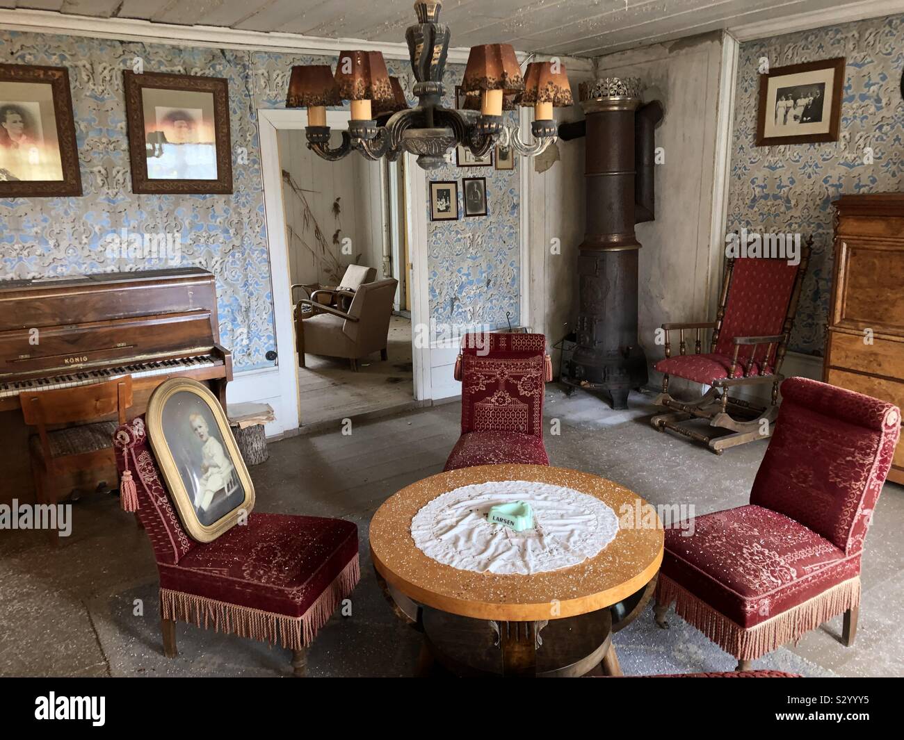 Decaying sitting room interior of old, abandoned farmhouse with vintage style furniture, a piano and framed portraits on walls with flock wallpaper. - Smartphone Captured Stock Image