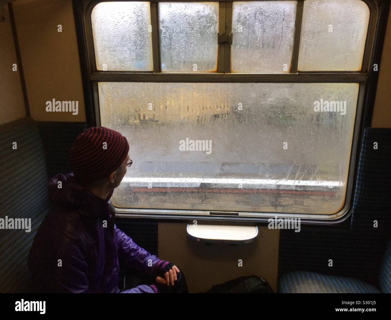 A man sits in an old fashioned train carriage looking out of the window on a cold rainy winters day - Smartphone Captured Stock Image