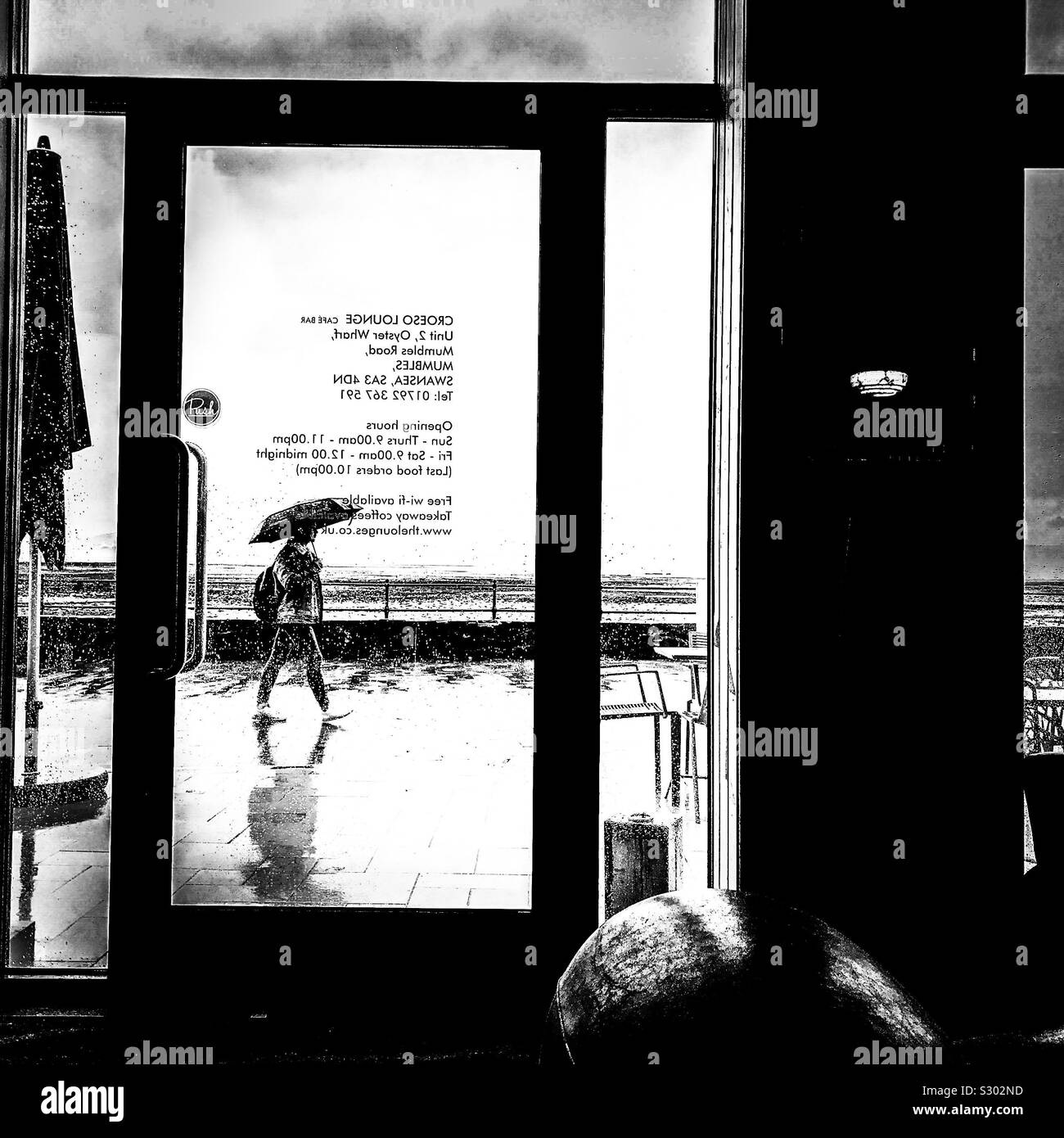 View out the glass window door of a modern cafe of a pedestrian walking passing by in the rain holding an umbrella by the seaside seafront promenade Stock Photo
