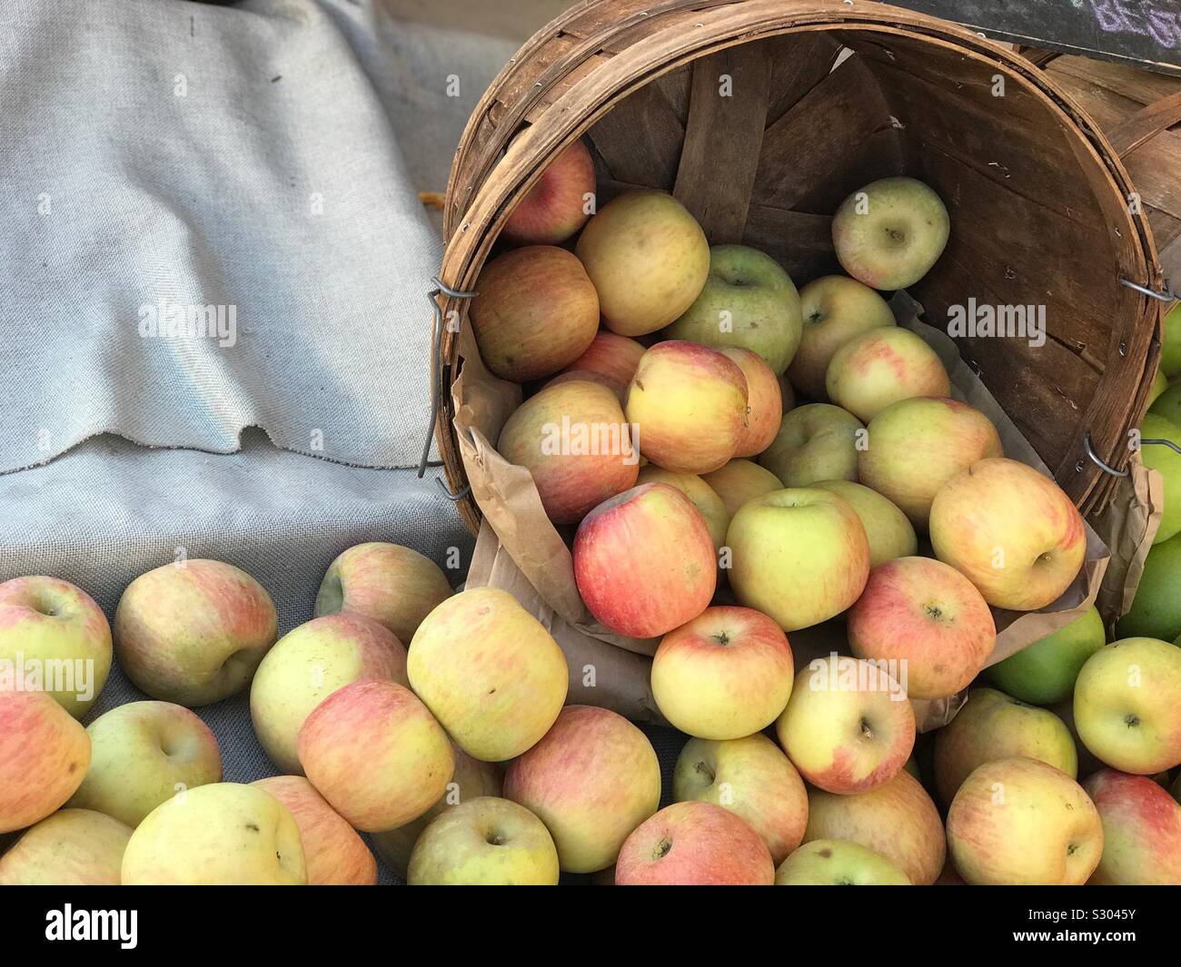 Ripe apples are shown spilling out of a basket in an exterior display at a farmers market. - Smartphone Captured Stock Image