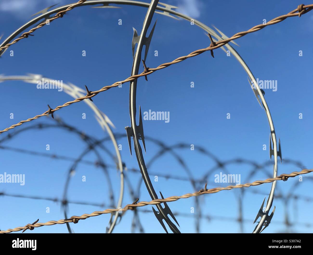Barbed and razor wire are shown up close, atop a fence, providing security for a secure area. - Smartphone Captured Stock Image