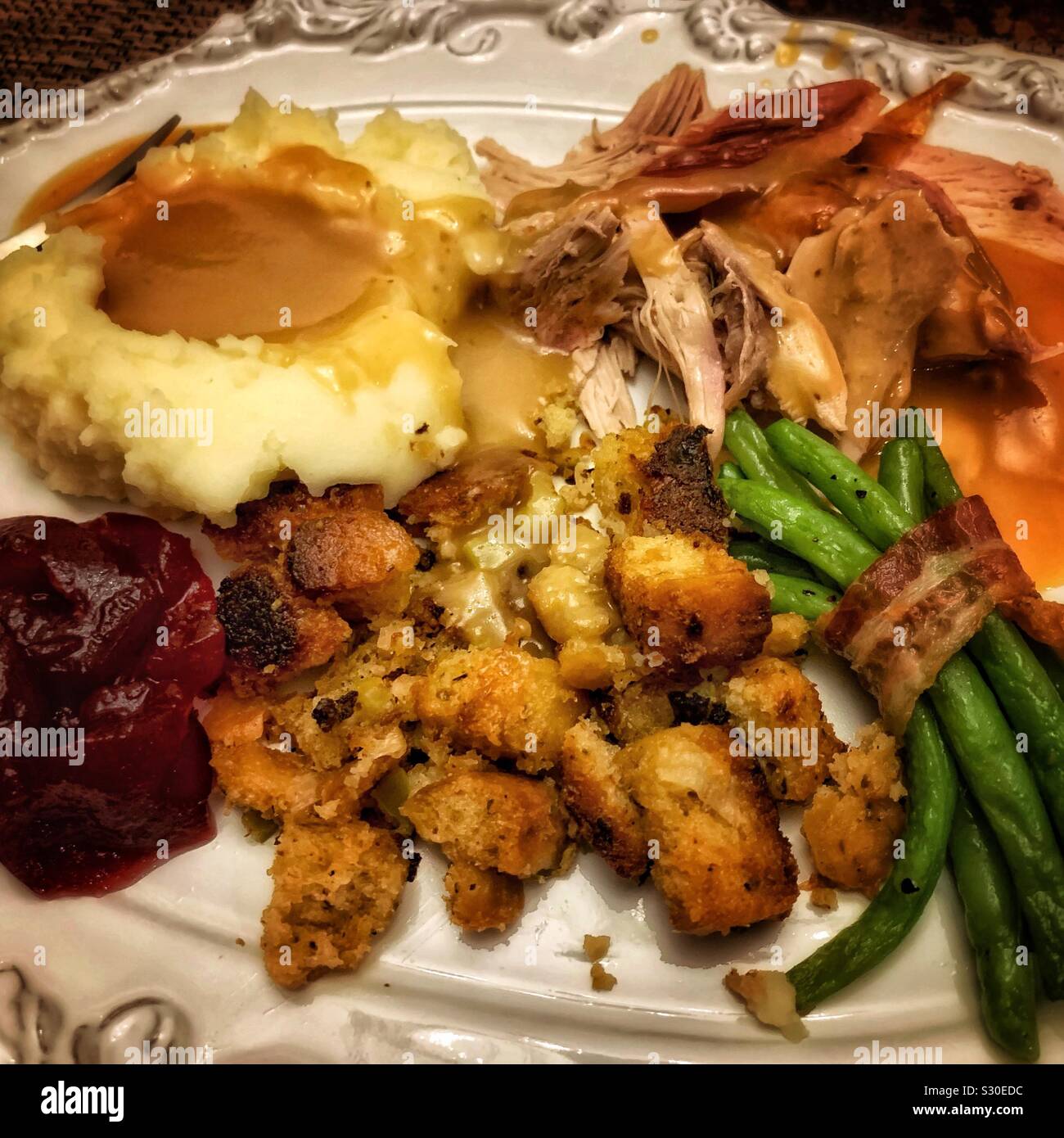 A Thanksgiving meal is plated featuring turkey, a bacon wrapped green bean bundle, dressing, cranberry sauce, and mashed potatoes with gravy. Stock Photo