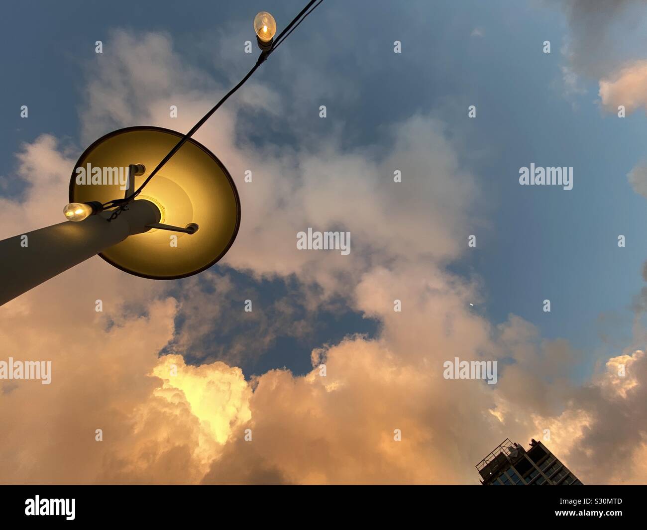 Looking up at lamp with amber glow and sunset, orange clouds in background. Top of a skyscraper can be seen in the corner. - Smartphone Captured Stock Image