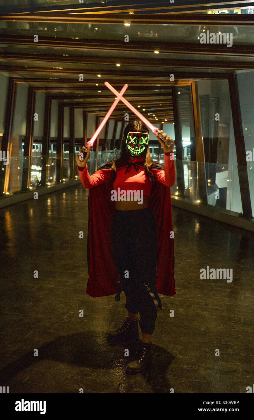 Green neon mask girl with red light sabers wearing cloak, in dark entrance - Smartphone Captured Stock Image