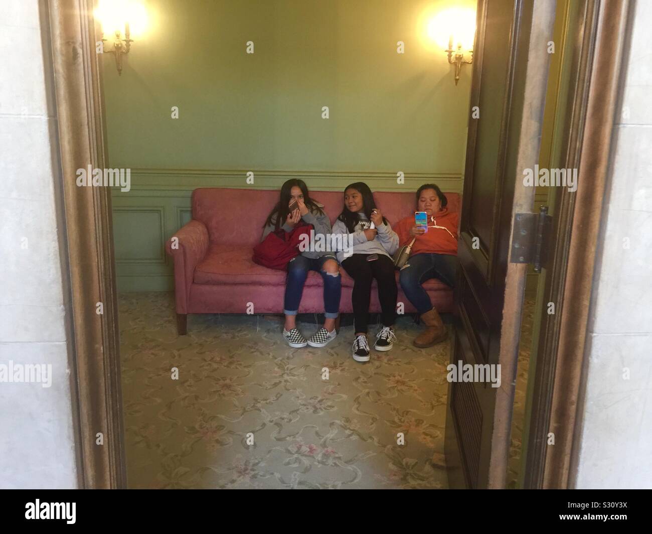 Girls on a pink couch holding cell phones in the bathroom lounge in the War Memorial Opera House during intermission of a San Francisco Opera matinee. San Francisco, California. Stock Photo