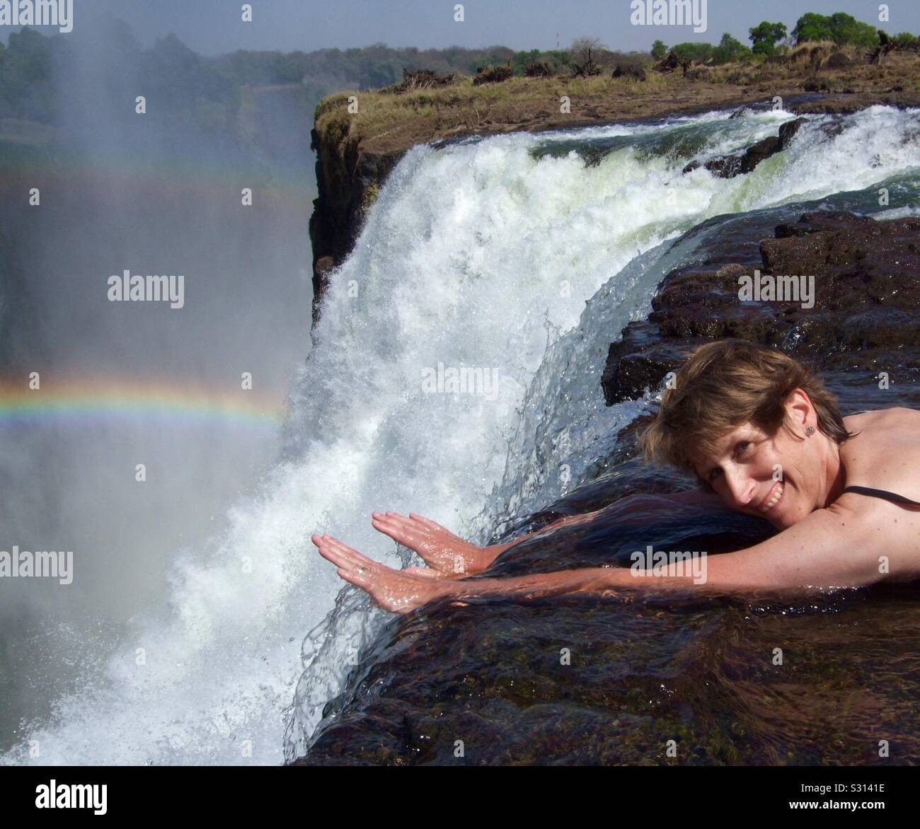 Woman lying in water at cliff edge of Victoria Falls at Devil’s Pool waterfall with a rainbow, Livingstone Island, Zambezi River, Zambia, Africa - Smartphone Captured Stock Image