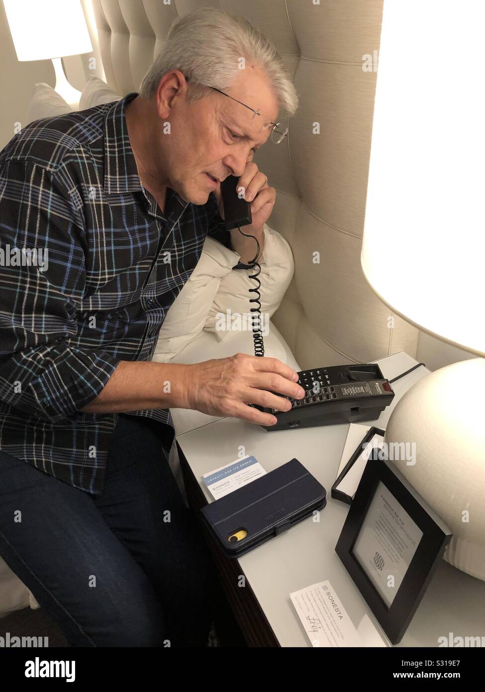 Man on a hotel room telephone making a call to the front desk with his cellular iphone on the table. - Smartphone Captured Stock Image