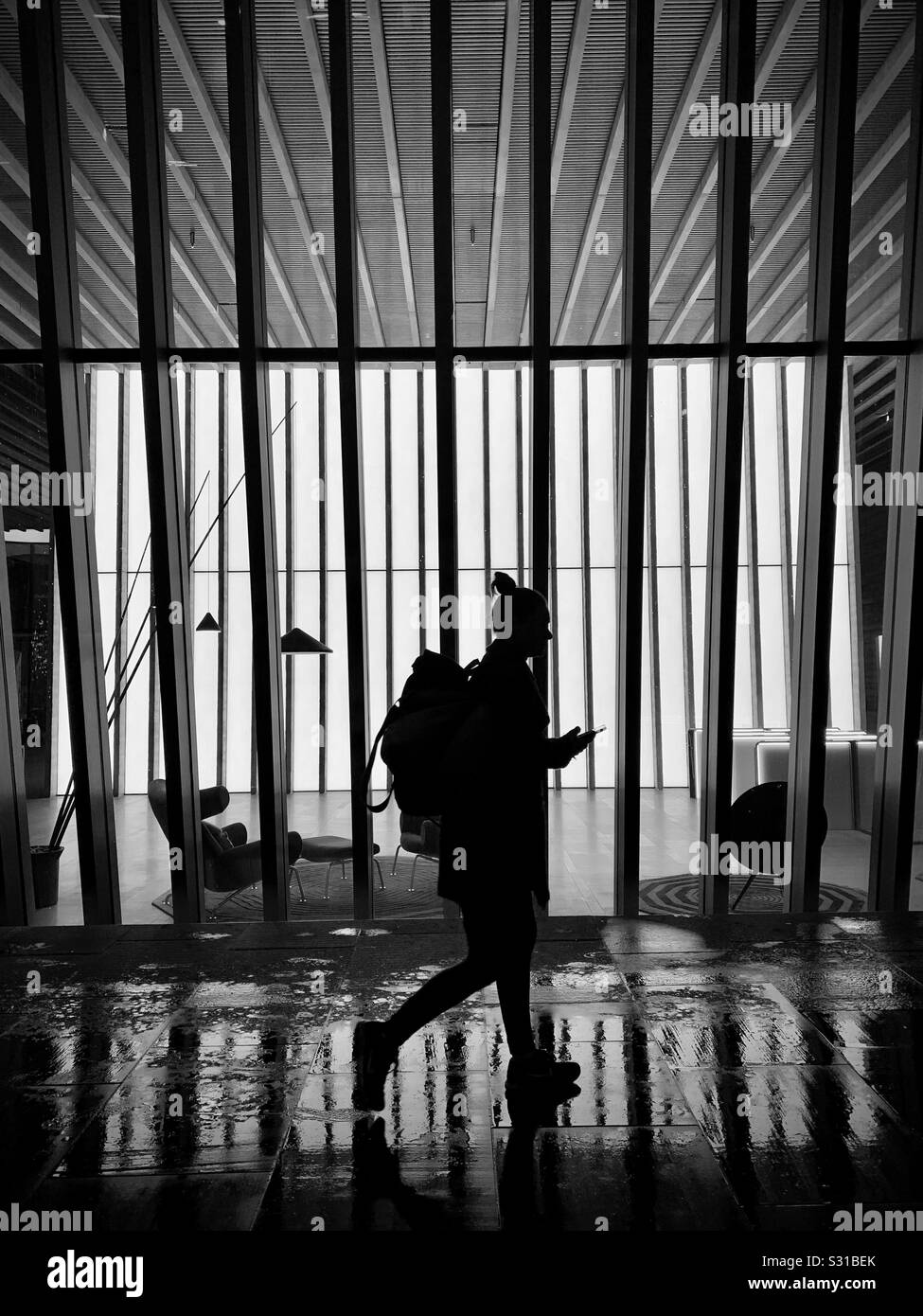 Silhouette of woman with backpack walking home and looking at phone with bright building in the background - Smartphone Captured Stock Image