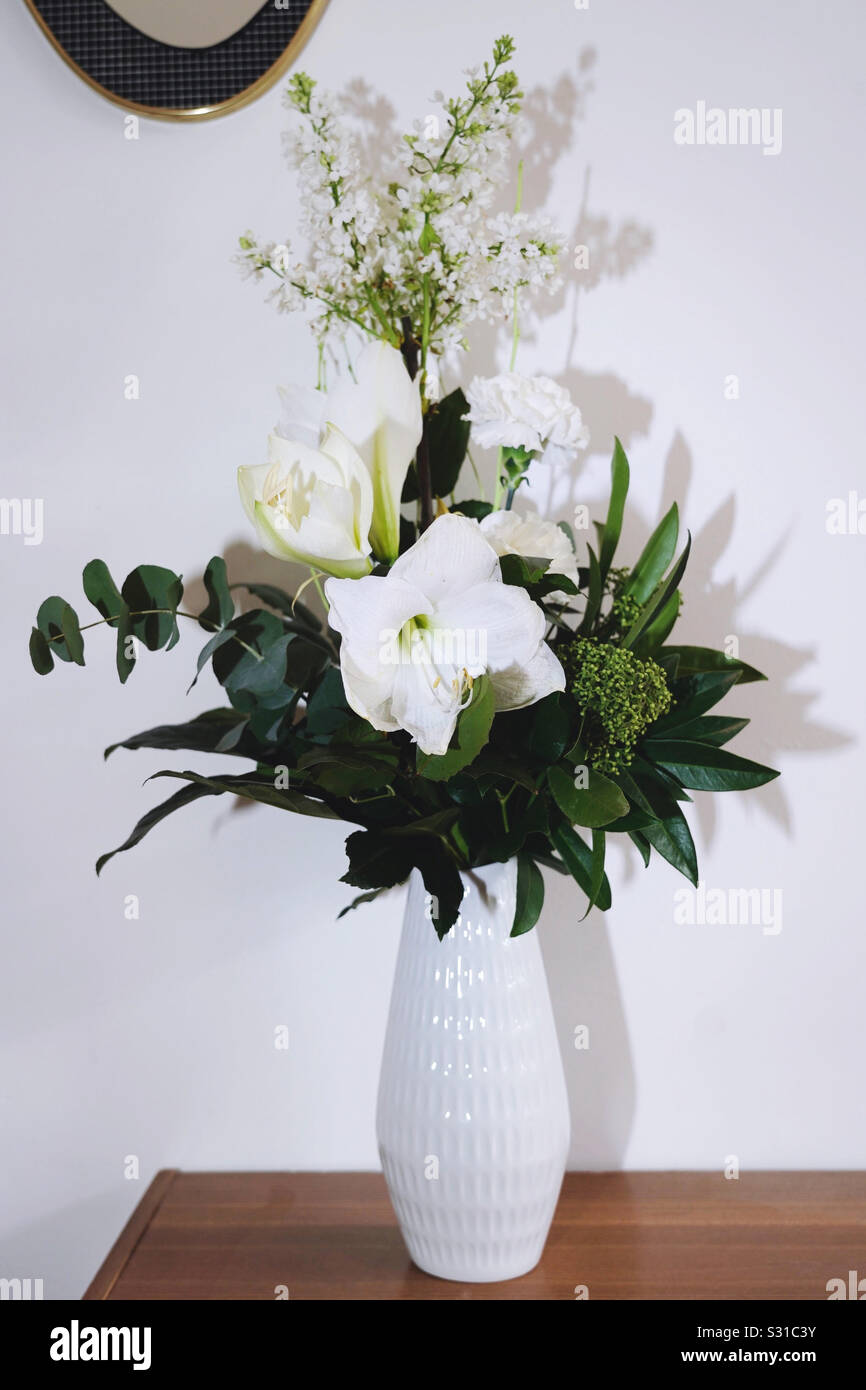 Bunch of flowers, white blossom and green leafs. In a white vintage porcelain vase on a teak shelf. Stock Photo