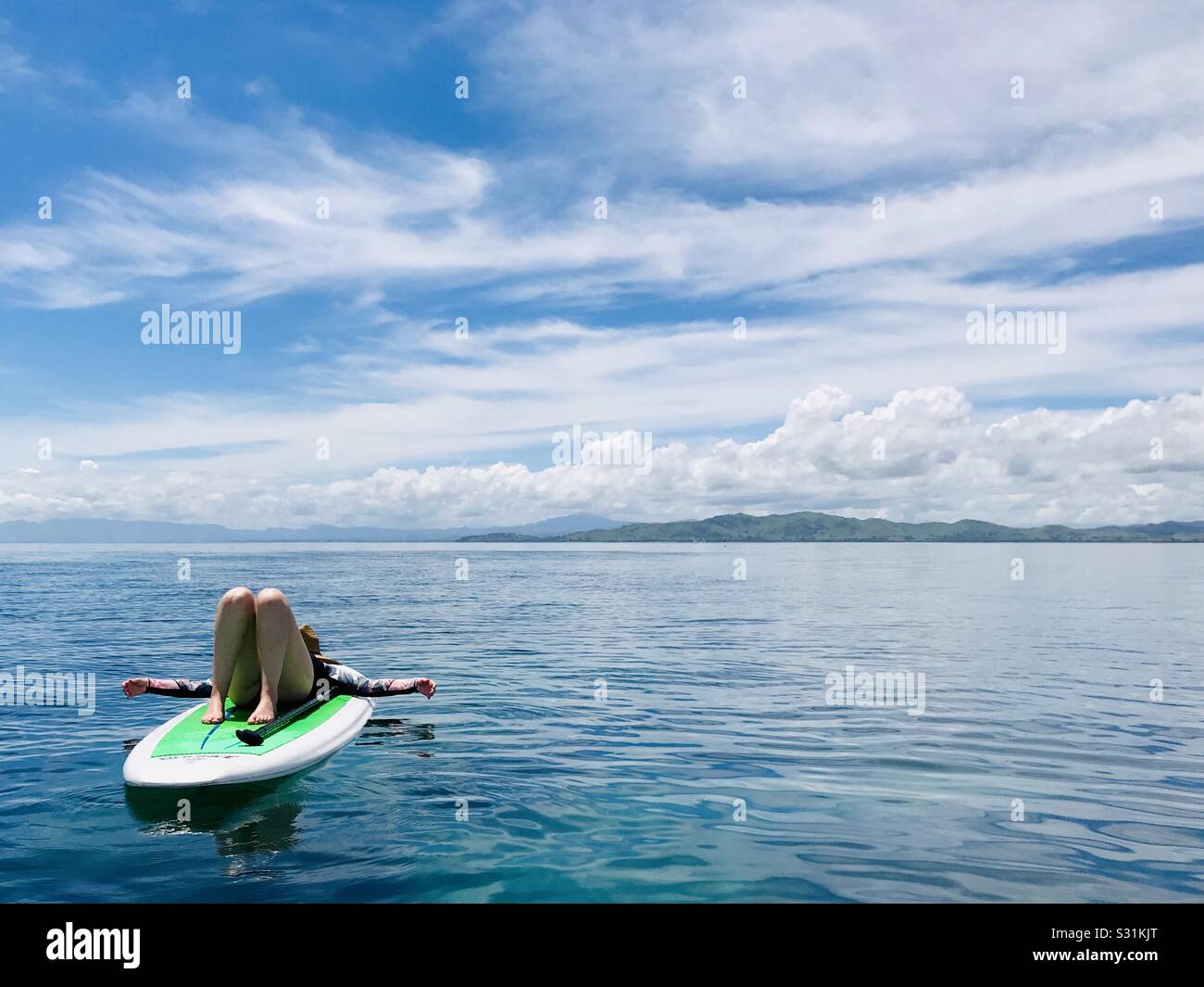A young female lays on a paddle board in crystal blue water. Tavarua Island, Fiji. Stock Photo