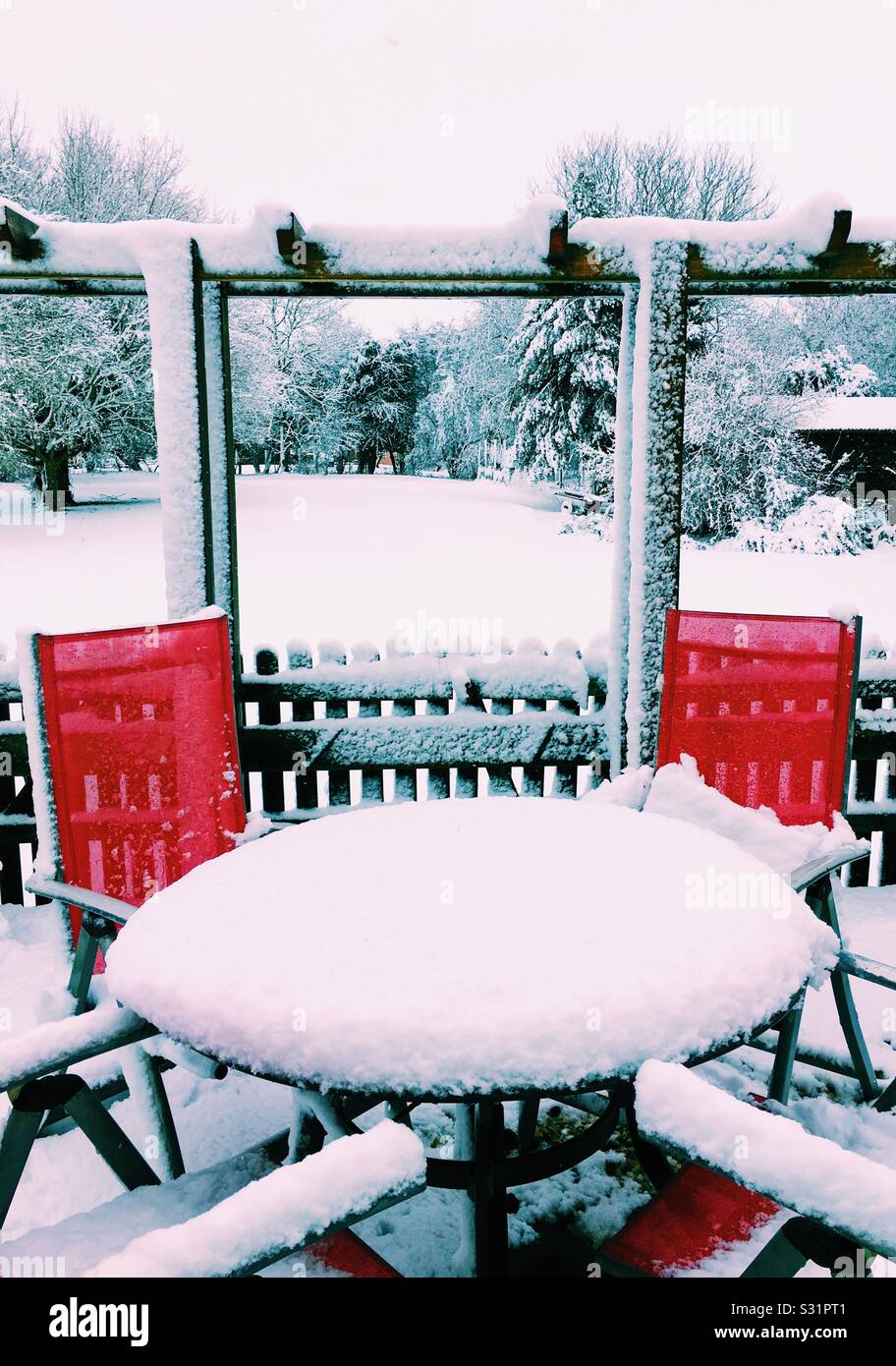 Table and red chairs covered in snow with large garden in background Stock Photo
