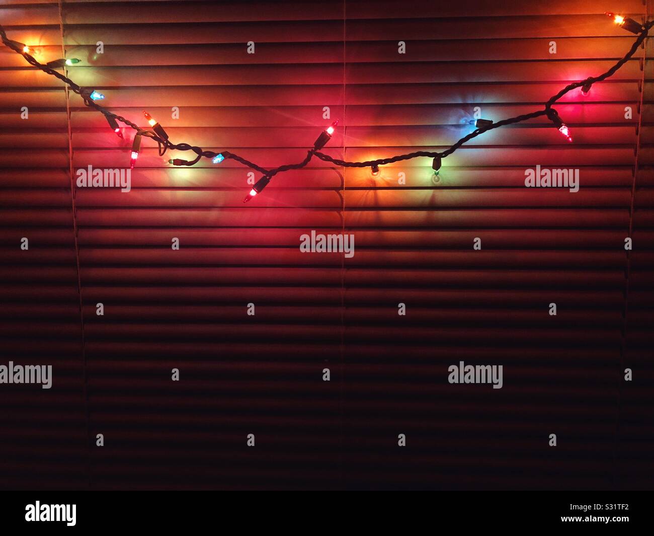 String of colorful christmas lights cast glow on window blinds - Smartphone Captured Stock Image