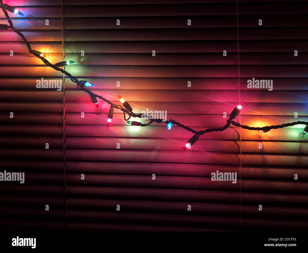 String of colorful christmas lights cast glow on window blinds - Smartphone Captured Stock Image