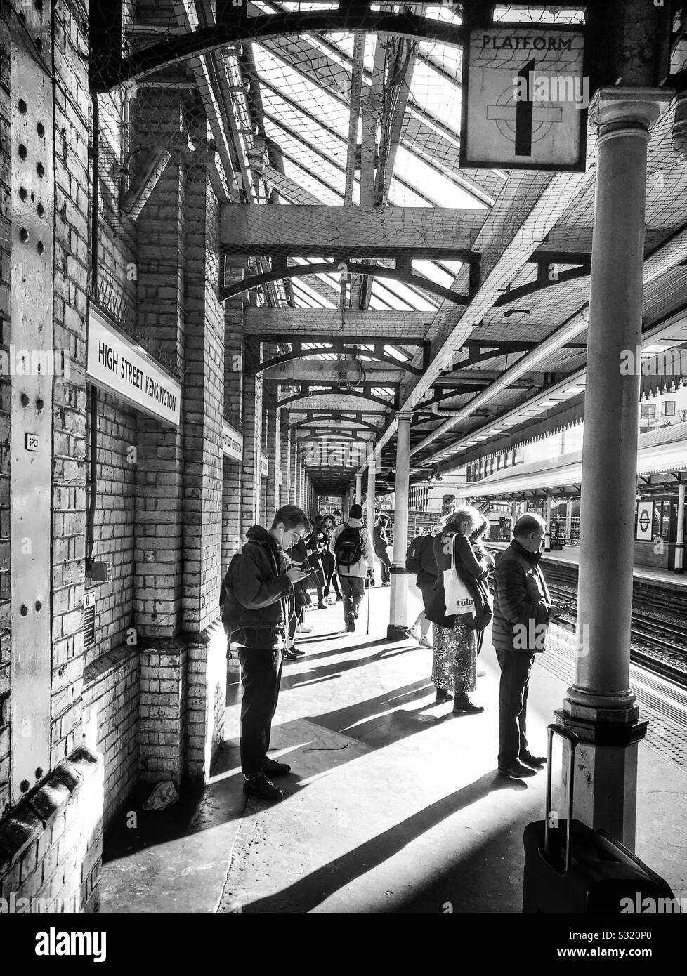Platform 1 of London Underground’s High Street Kensington Station, with passengers waiting for the next train. Bright sunlight casts long shadows of this scene, captured in black and white. - Smartphone Captured Stock Image