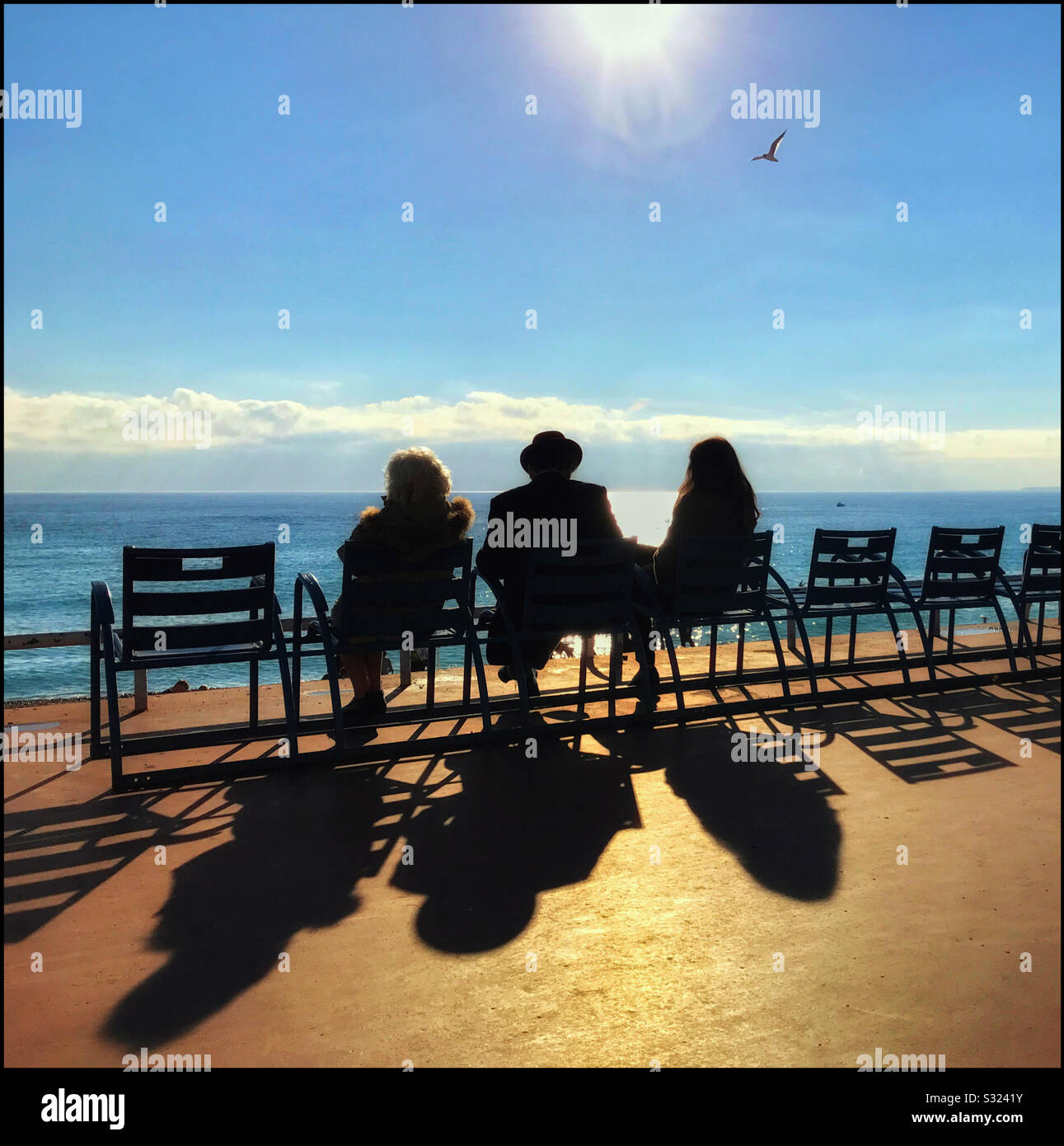A man and 2 women sit on chairs in the bright sunshine watching the sun start to set over the Mediterranean Sea. Interesting shadows and a well placed seagull! Photo © COLIN HOSKINS. - Smartphone Captured Stock Image