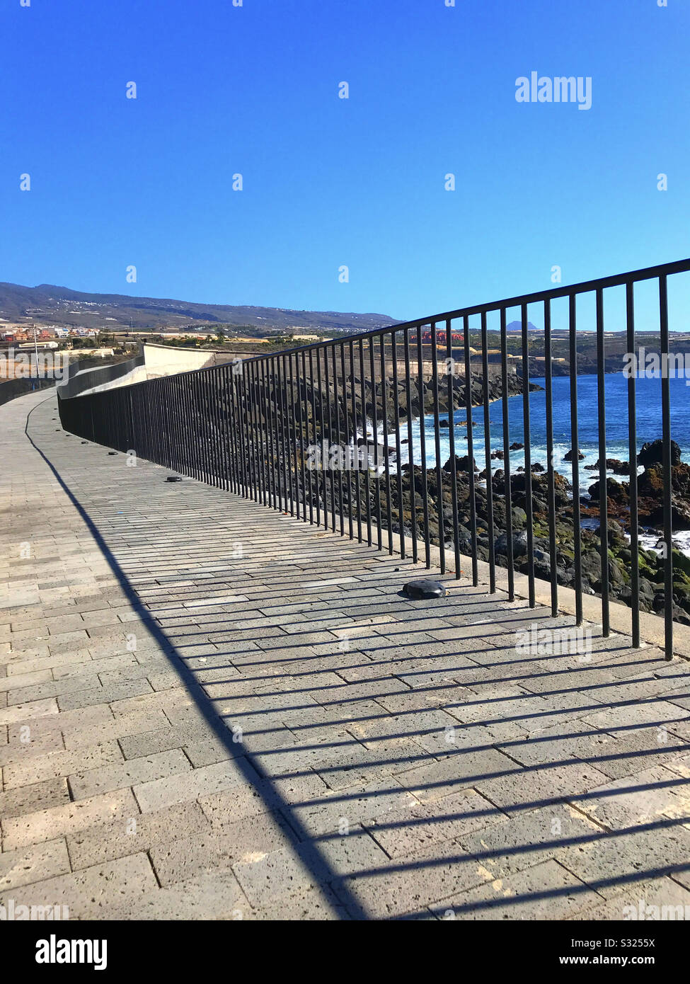 Fence rails on the promenade disappear to a vanishing point and throw shadows on the pavement at the coast in Playa San Juan Tenerife - Smartphone Captured Stock Image