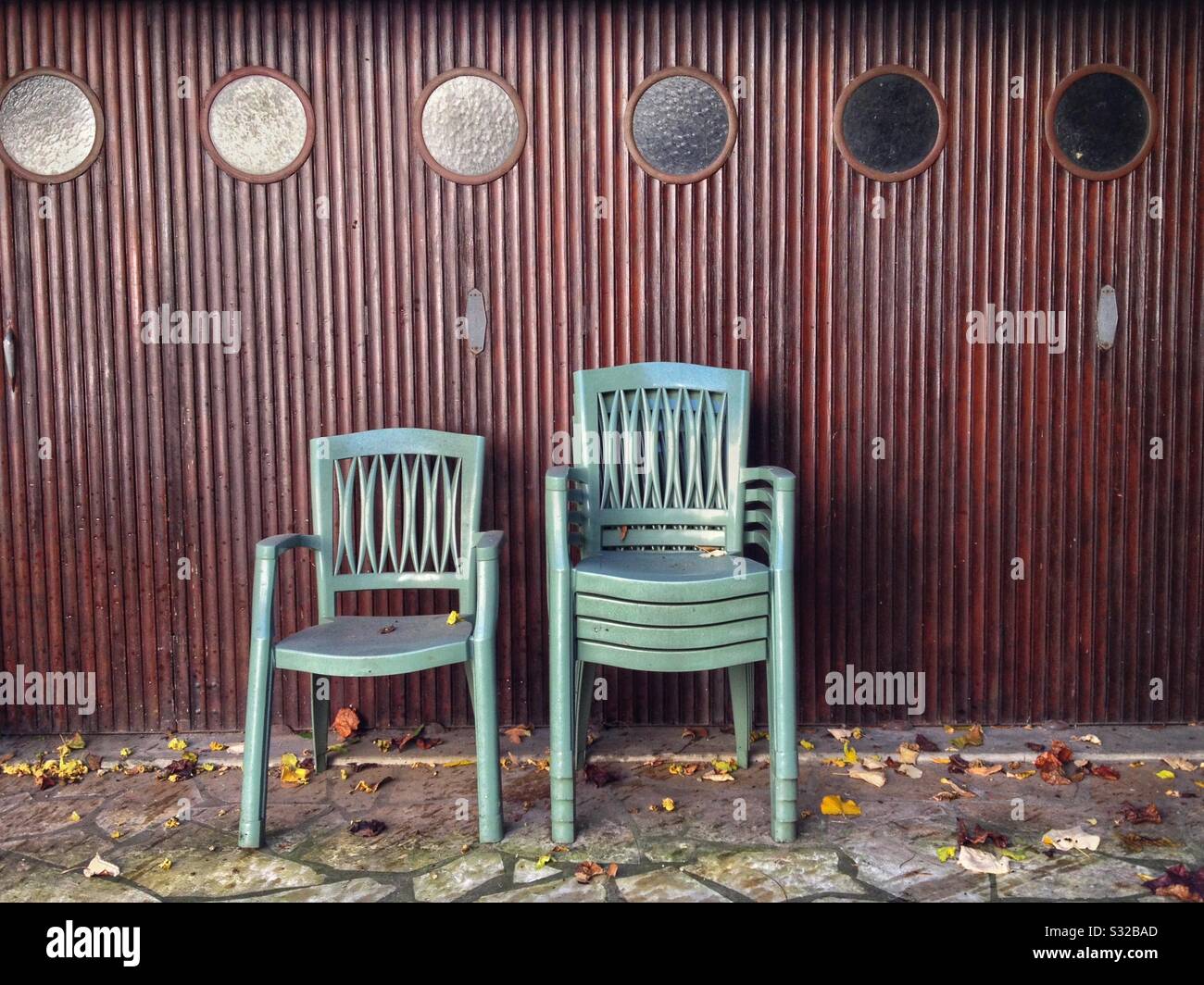 Garden chairs stacked up against a garage door with small circular windows. - Smartphone Captured Stock Image