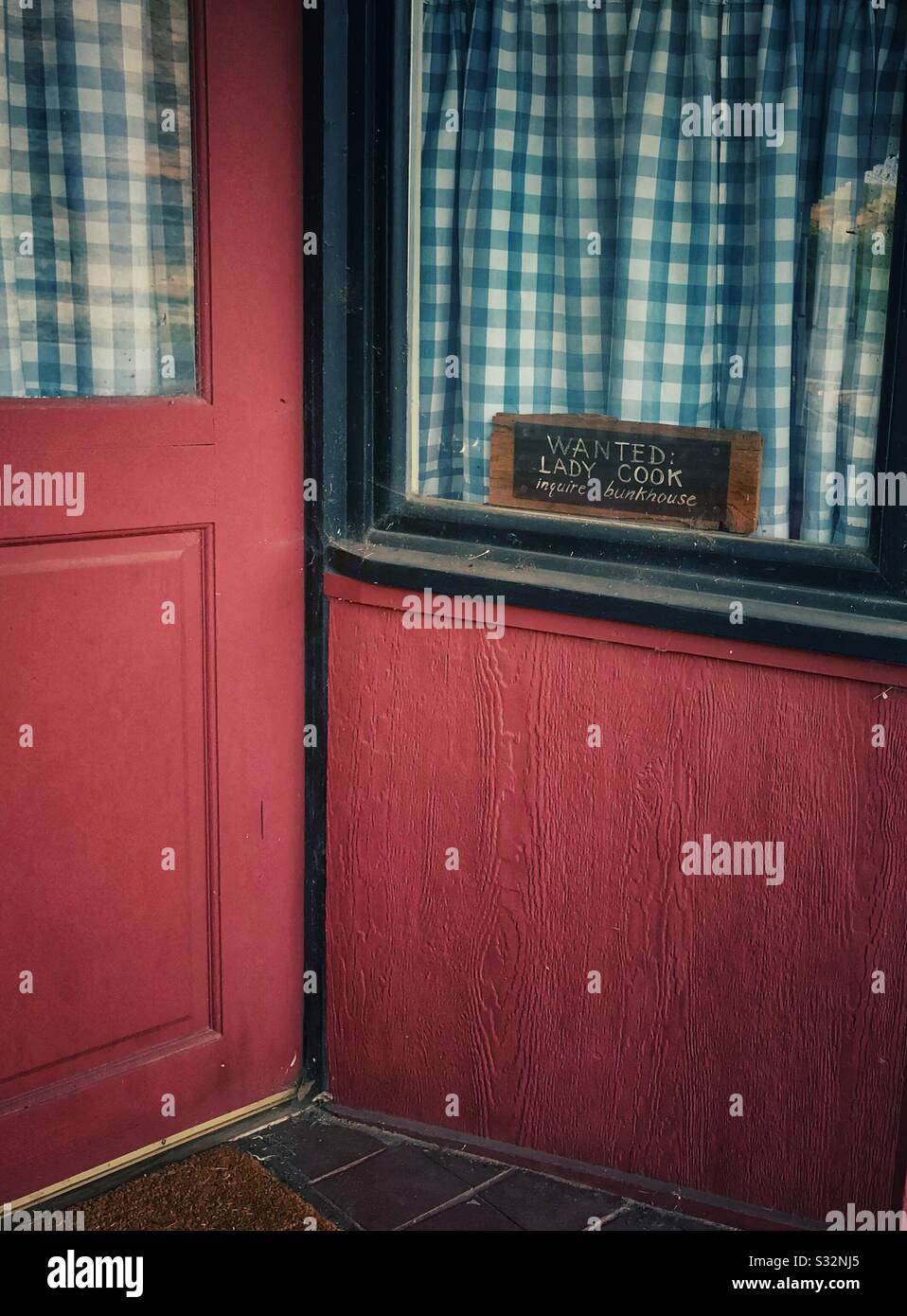 Red door and blue gingham curtains at restaurant with sign in window- Wanted: Lady Cook Inquire Bunkhouse Stock Photo