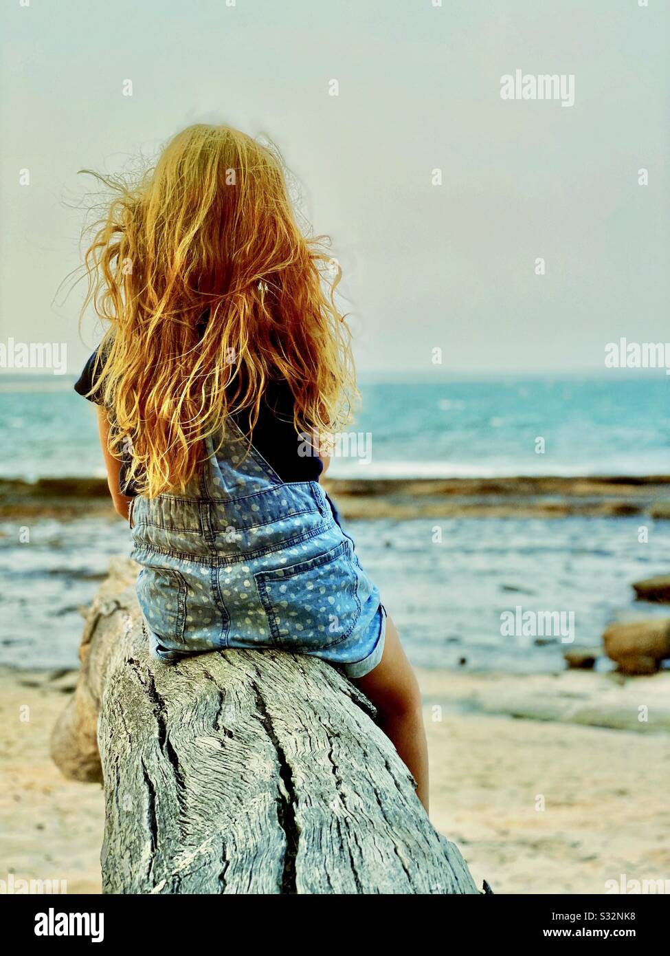 Back view of young girl with long red hair seating on log looking to sea. Concepts of dreaming, wanderlust, childhood and lifestyle Stock Photo