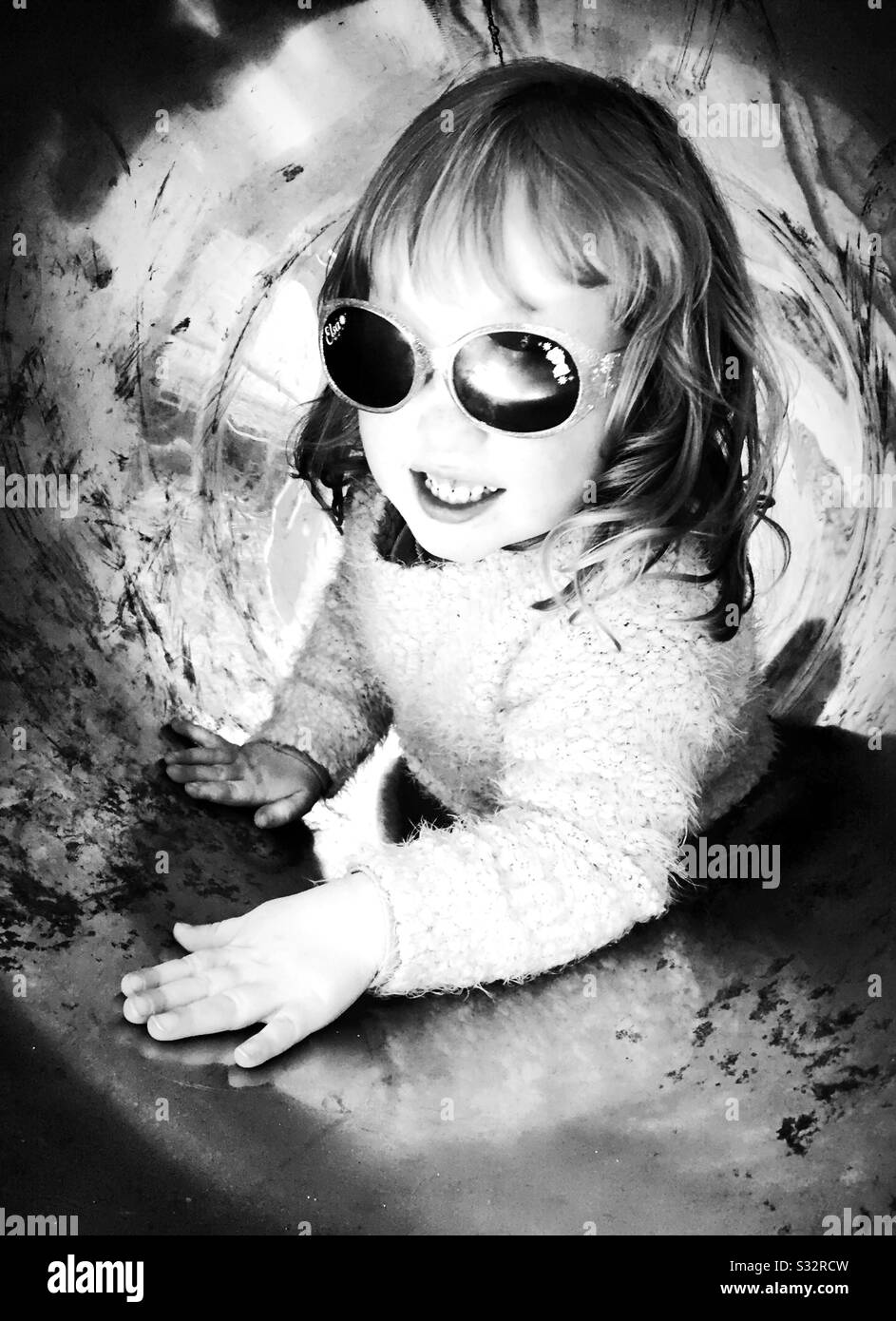 Young girl wearing sunglasses in metal play tunnel. Black and white Stock Photo