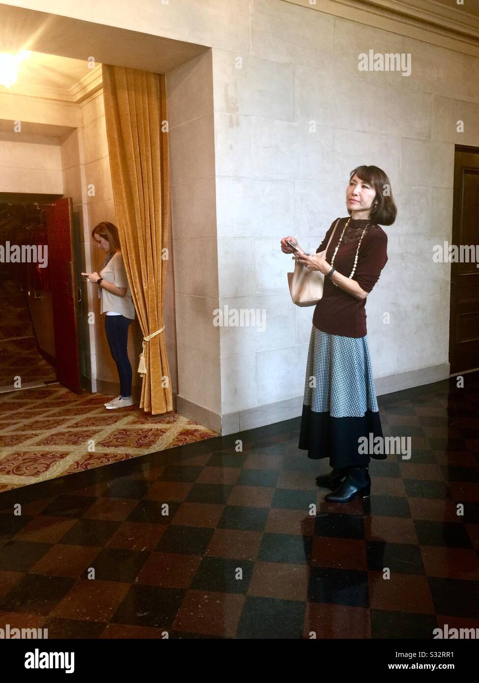 Two women waiting in the halls at the elegant War Memorial Opera House during intermission. One woman waiting for a friend, the other looking at her cell phone. San Francisco, California. Stock Photo