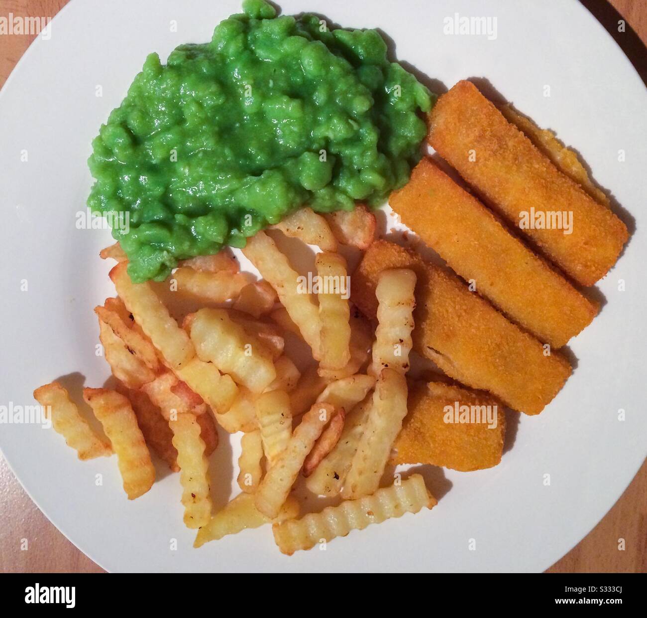 A complete meal of fish fingers, mushy peas and chips, on a white plate, viewed from above. Stock Photo
