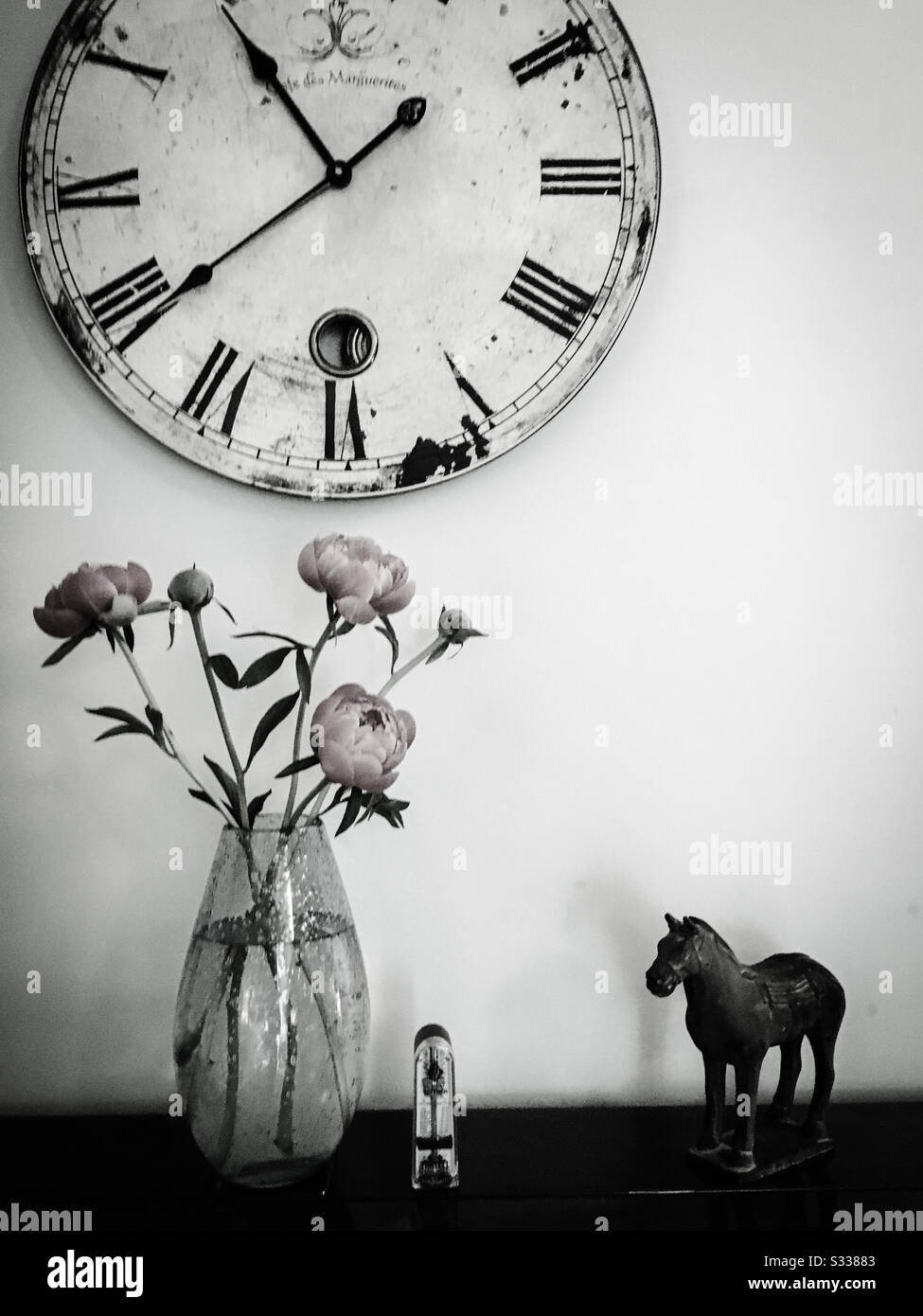 A black and white still life photograph of a clock, oriental horse ornament, metronome and a vase of peony flowers, all placed on a piano. Stock Photo
