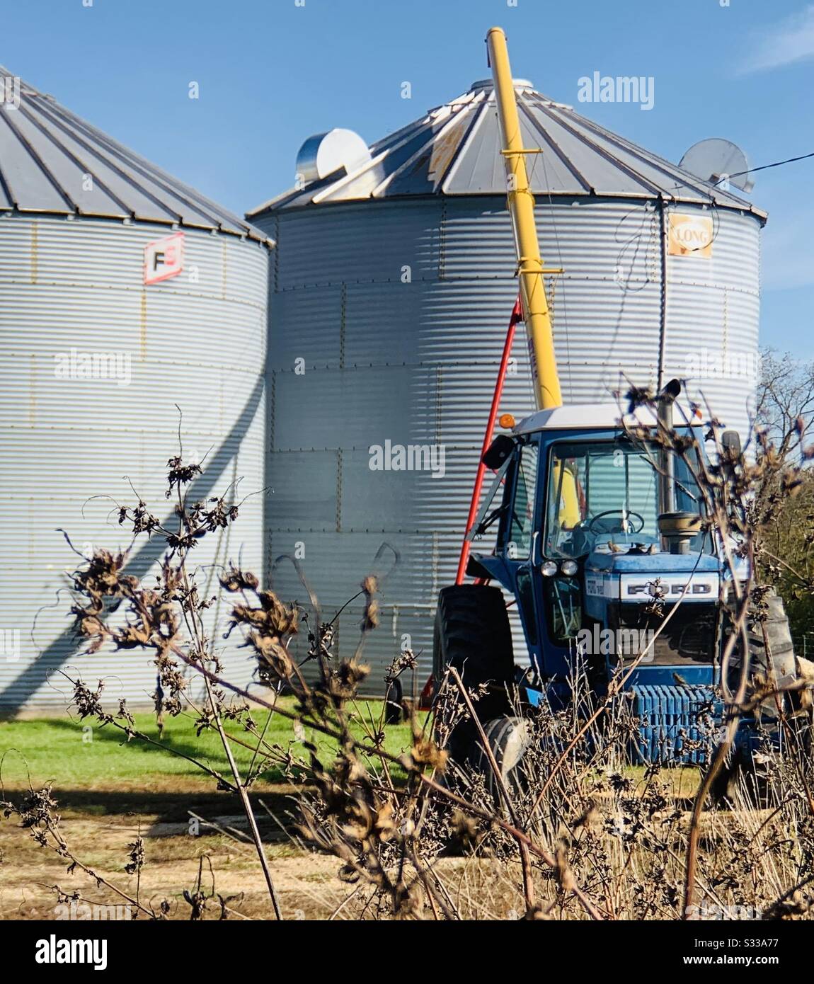 DUBUQUE, IOWA, Fall 2019--Landscape photo of retro blue and white Ford tractor at work in front of grain bins under a bright blue sky on fall harvest day. Stock Photo