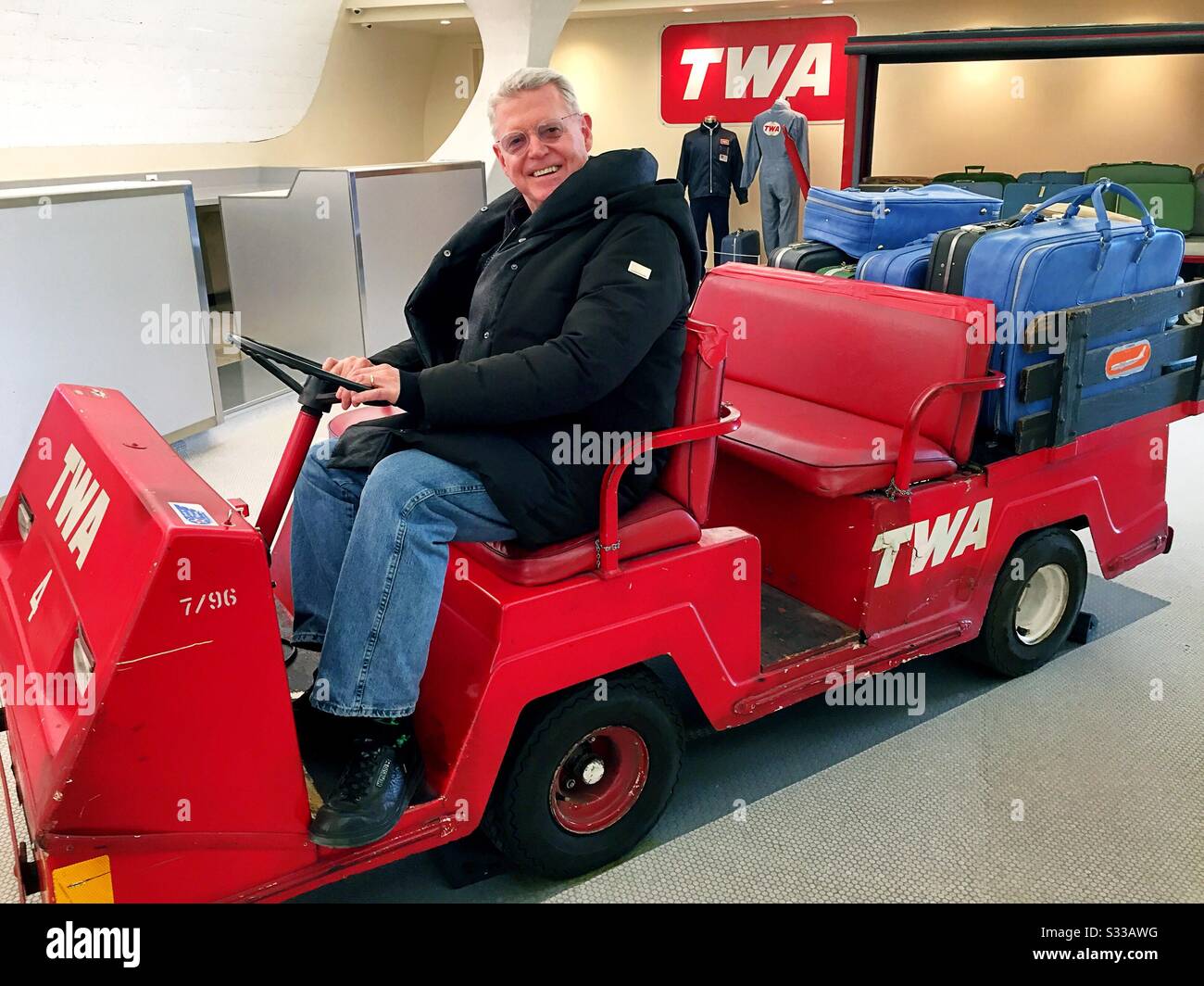 Senior hotel guest poses on a vintage luggage cart in the lobby of the TWA hotel, JFK airport, NYC, USA - Smartphone Captured Stock Image