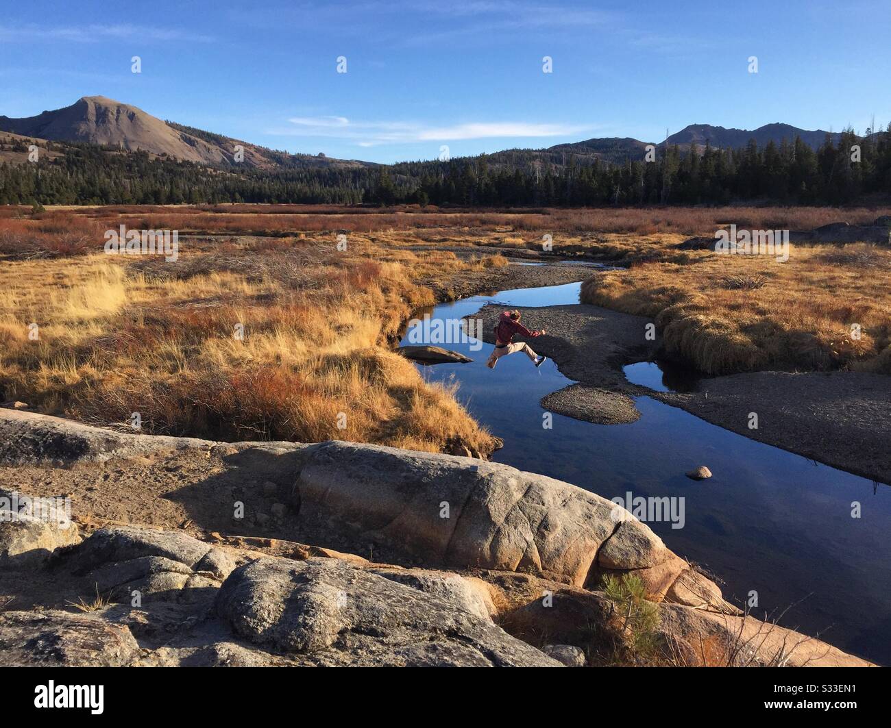 A boy jumps across a river winding through a valley with mountains in the distance. Stock Photo