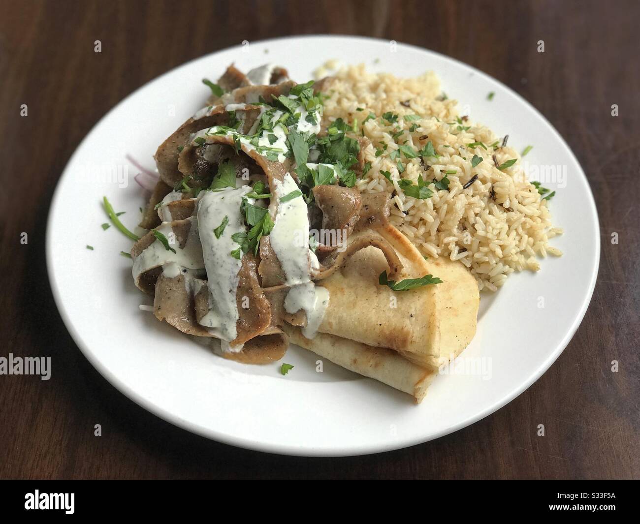 A beef and lamb gyro plate, consisting of meat covered with tzatziki sauce, rice, and pita bread, is shown on a plate at a Greek restaurant. - Smartphone Captured Stock Image