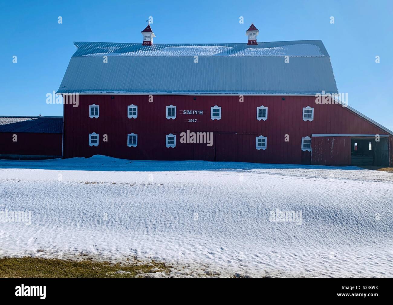 DUBUQUE, IOWA, 02/23/20–Landscape scenic photo of beautiful red barn with white framed windows with snow covered roof under a bright blue sky on a sunny day. Stock Photo