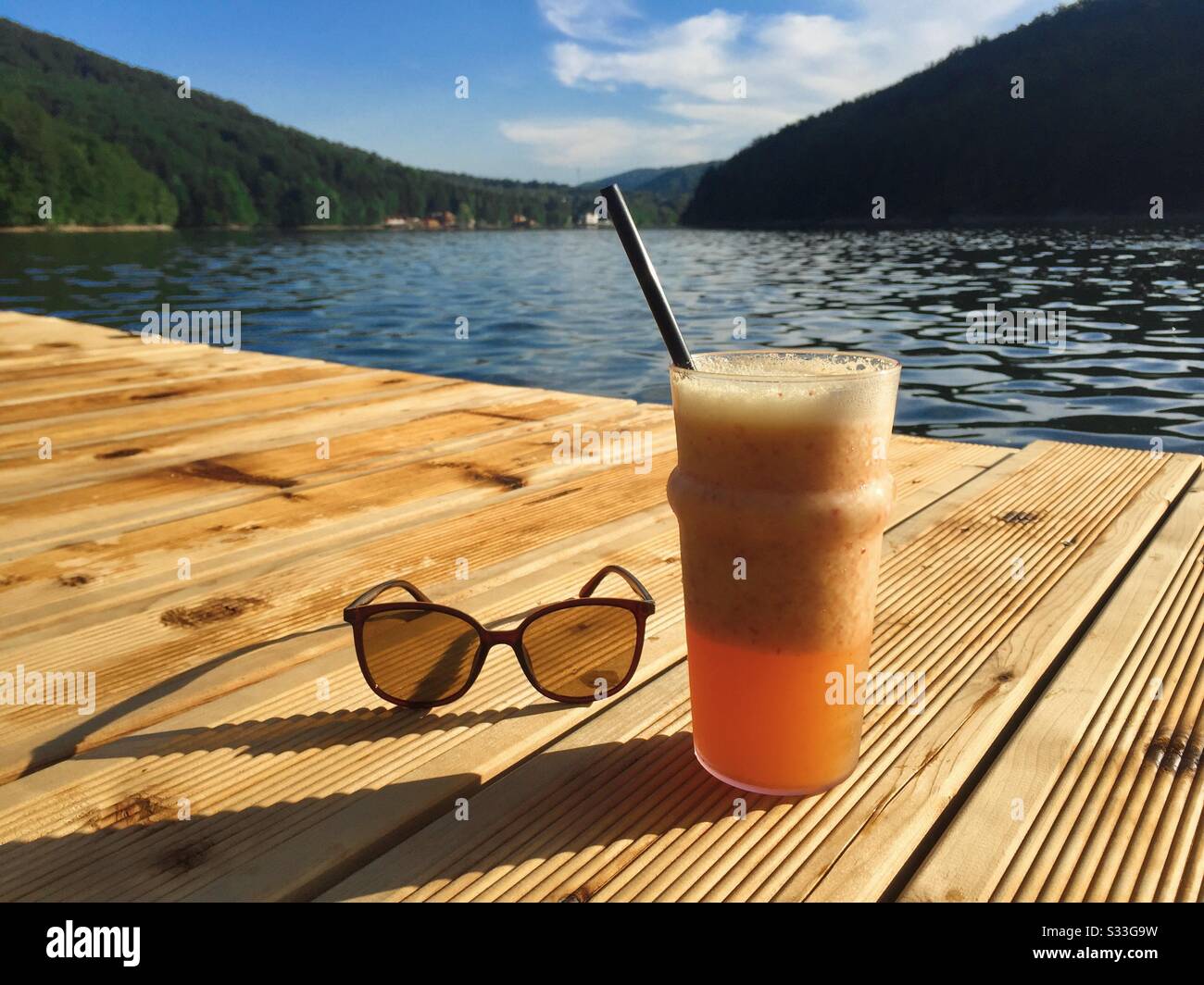 Glass of juice and a pair of sunglasses on wooden pontoon near the lake - Smartphone Captured Stock Image