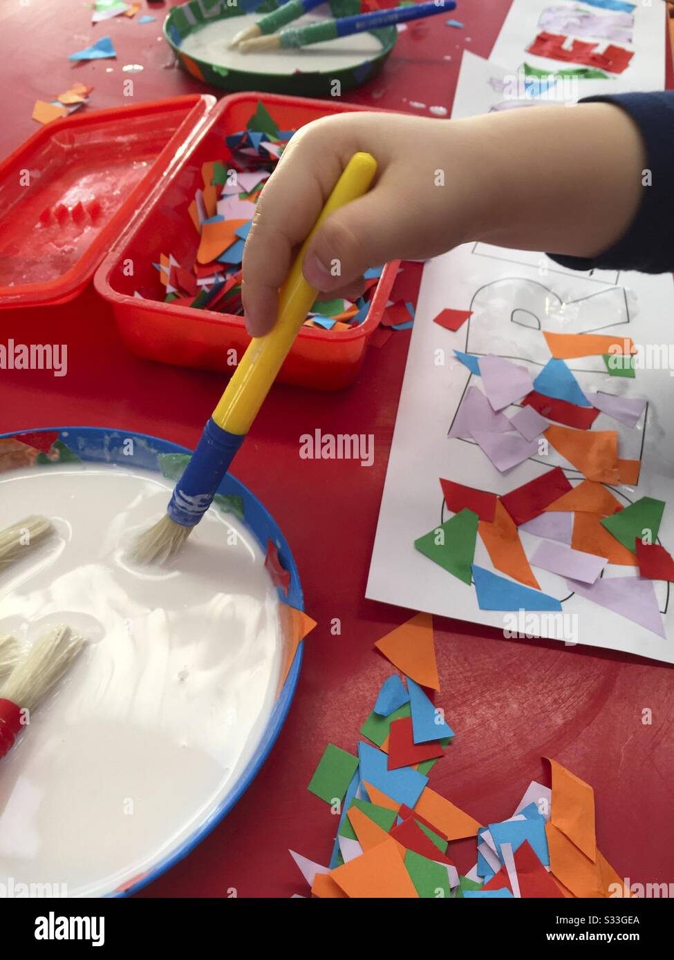 Child’s Hand with Brush Gluing Coloured Shapes onto Pieces of Paper - Smartphone Captured Stock Image